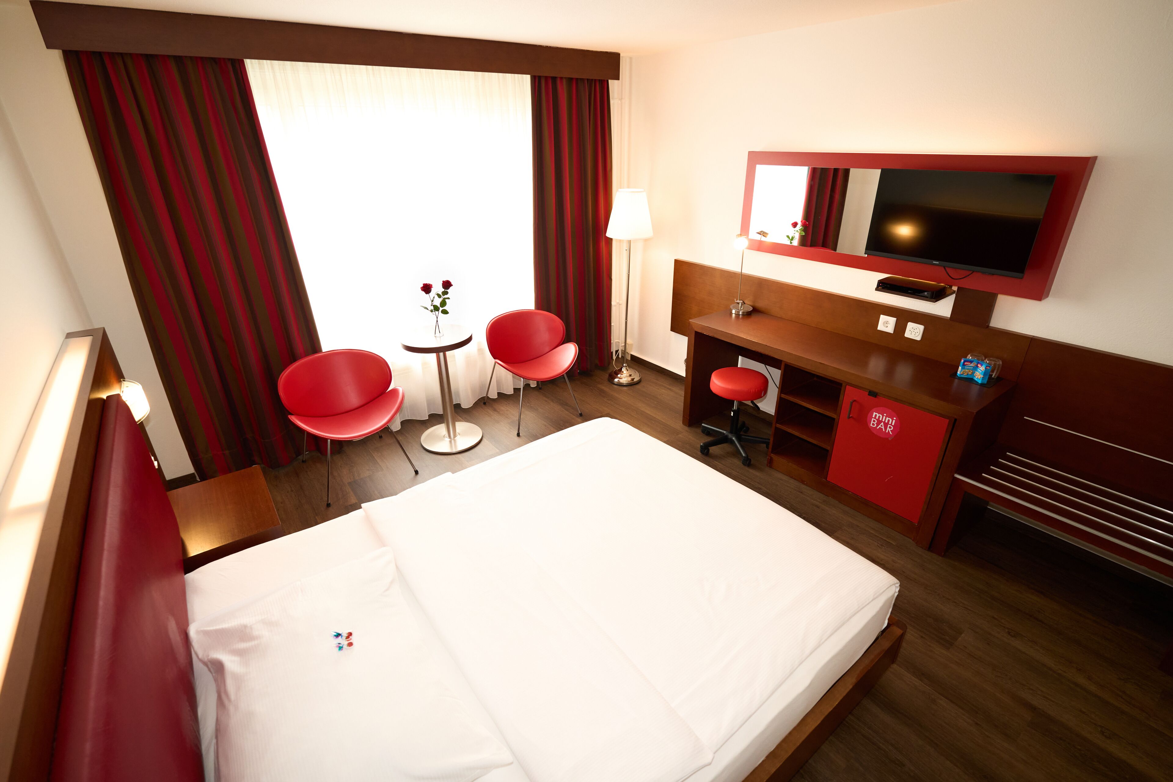 Une chambre d’hôtel avec un lit, deux fauteuils rouges, une petite table avec des fleurs, un bureau avec une chaise, un téléviseur fixé au mur et un mini-bar.