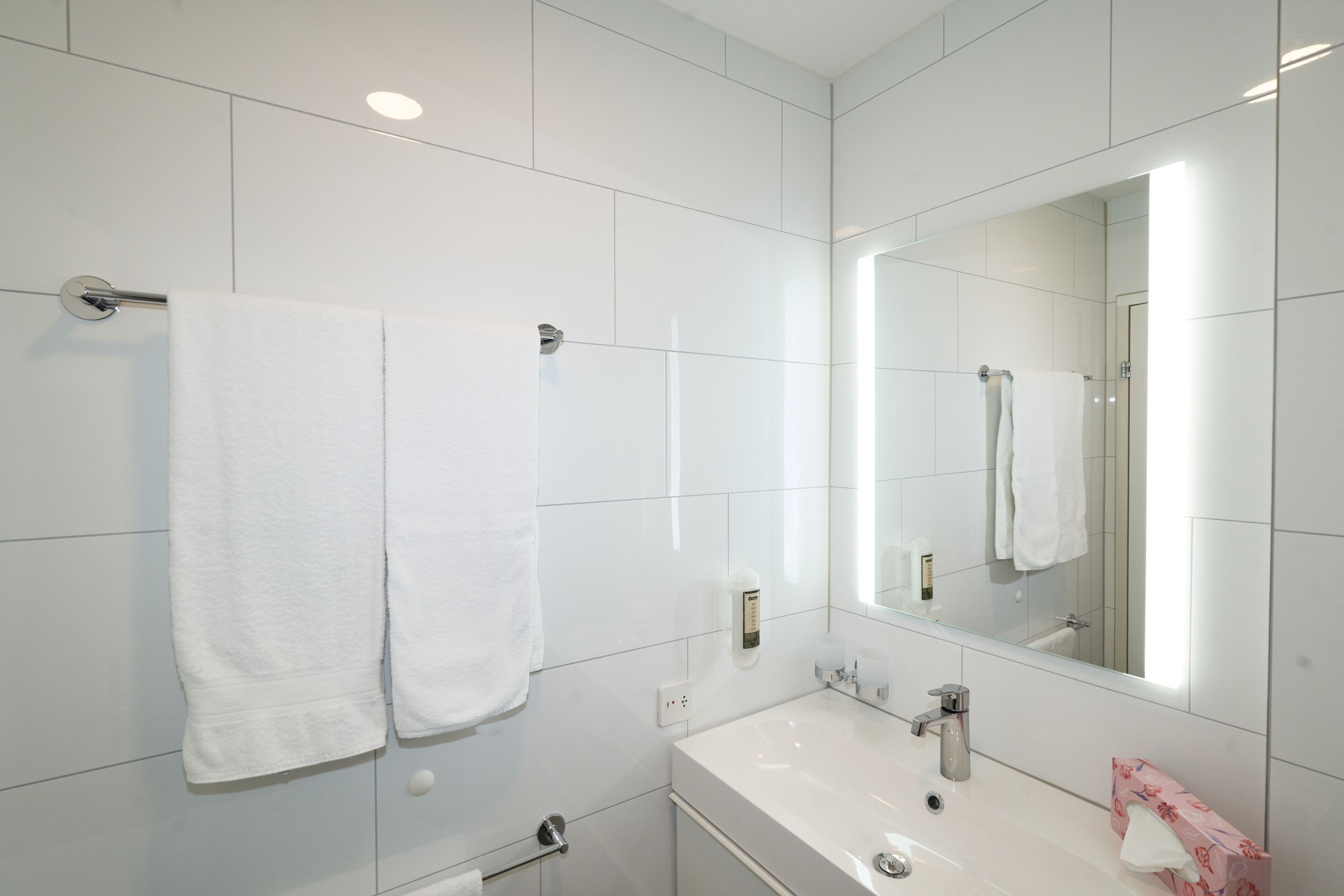 A modern bathroom with white tiles, a large mirror, a sink, and two towels hanging on a towel rack.