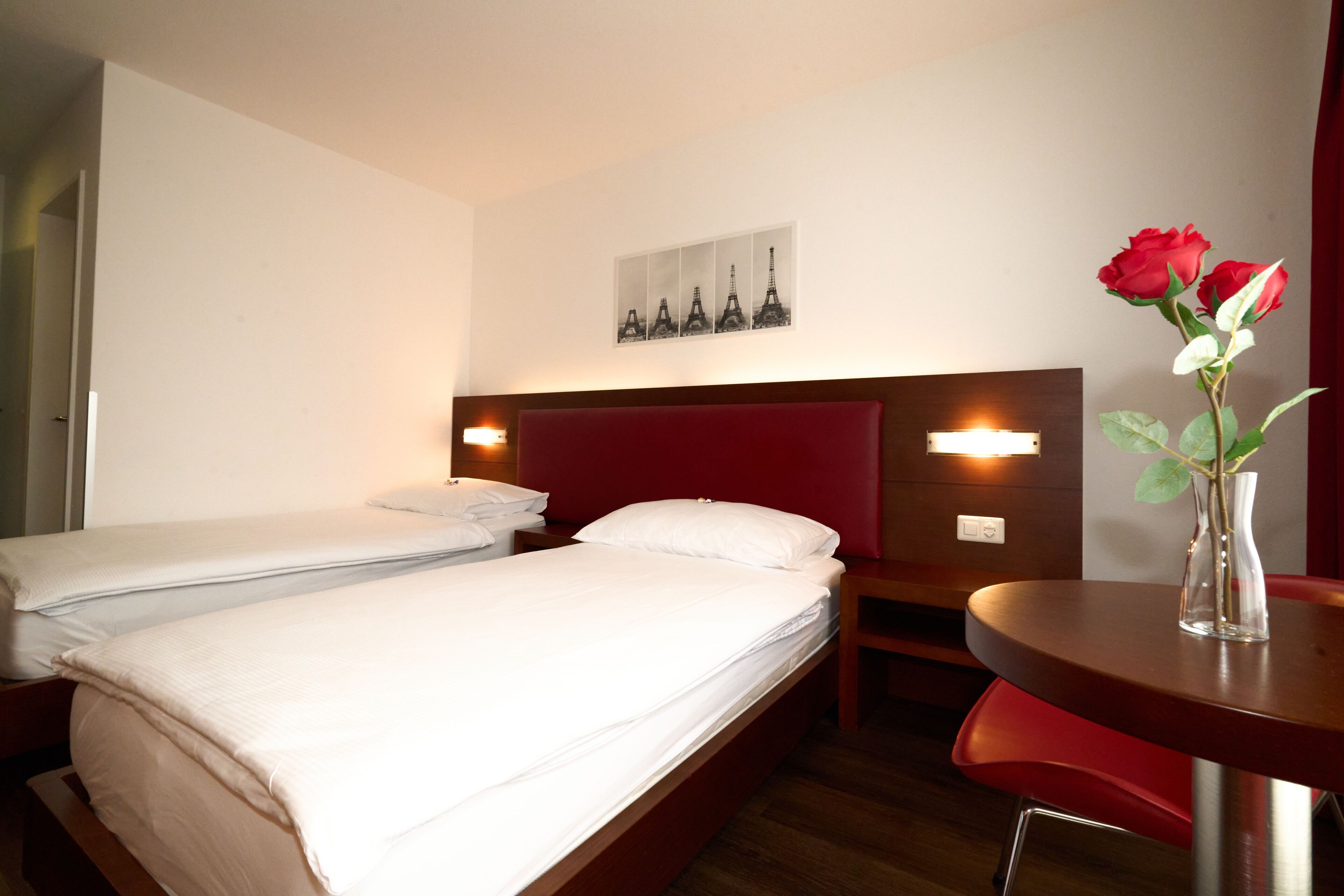 A hotel room with two single beds, a wooden headboard, a red chair, a small round table with a vase of flowers, and a wall-mounted picture of the Eiffel Tower.
