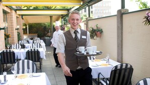 A waiter carrying a tray with cups and a small plate, serving guests at an outdoor dining area with tables and chairs.