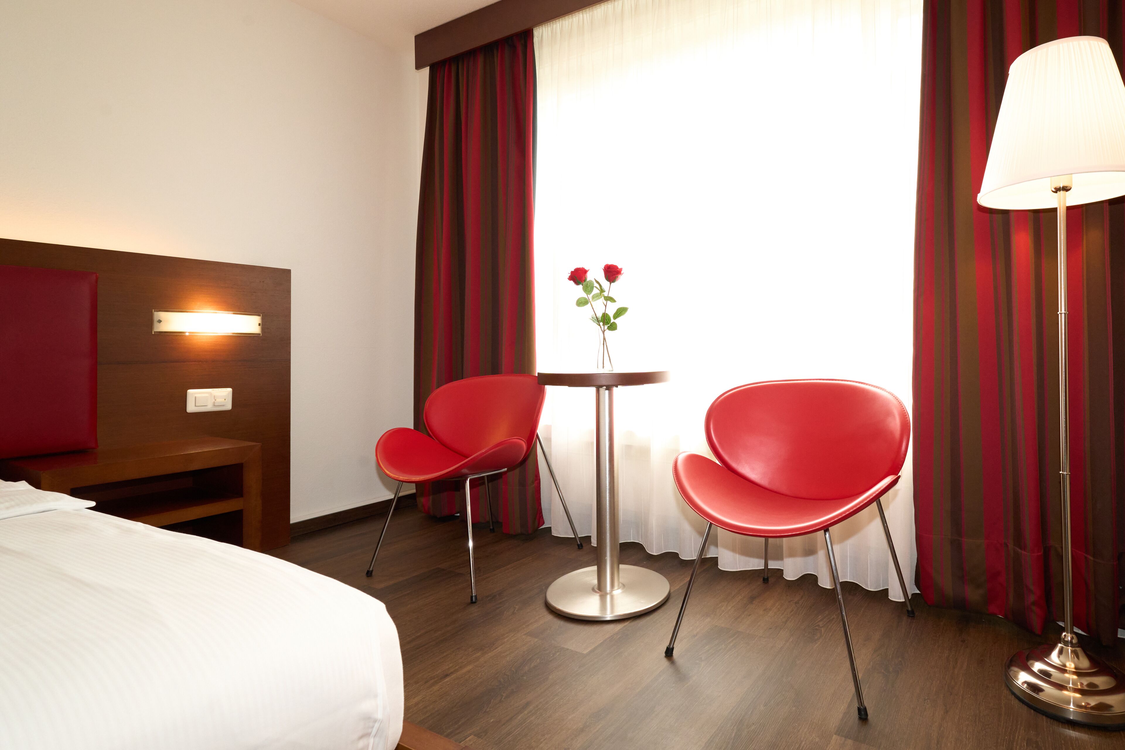 Une chambre d’hôtel avec un lit, une petite table et deux chaises rouges, un vase avec des fleurs et une lampe.