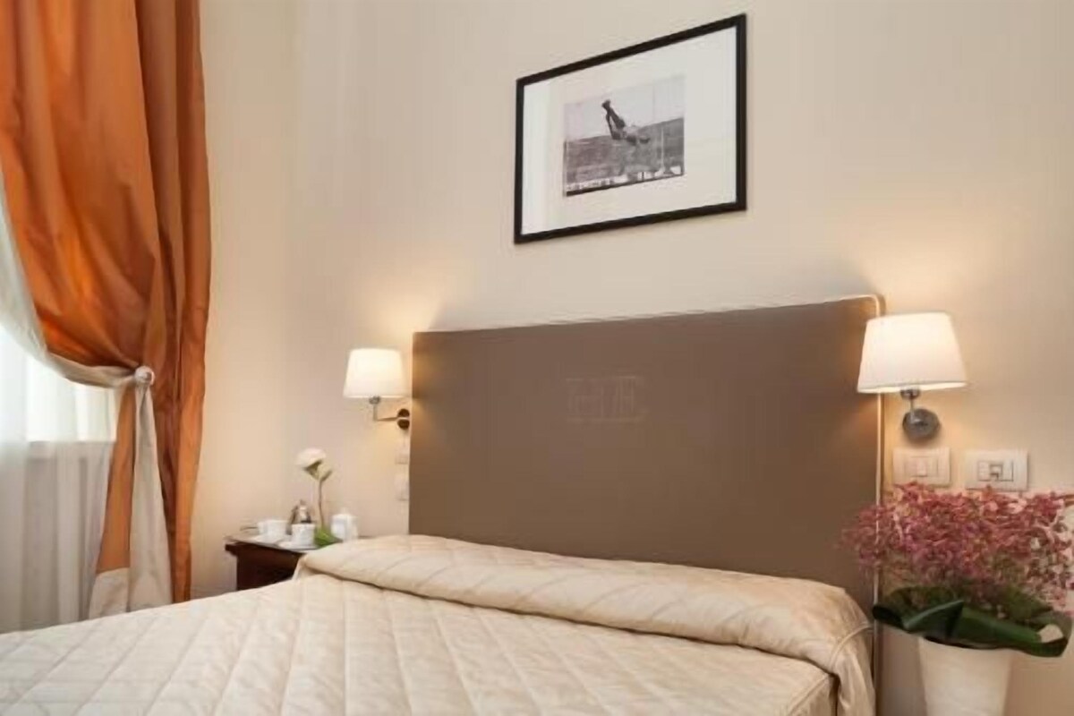 A hotel room with a large bed, two bedside lamps, a framed picture on the wall, and a nightstand with a lamp and a vase of flowers.