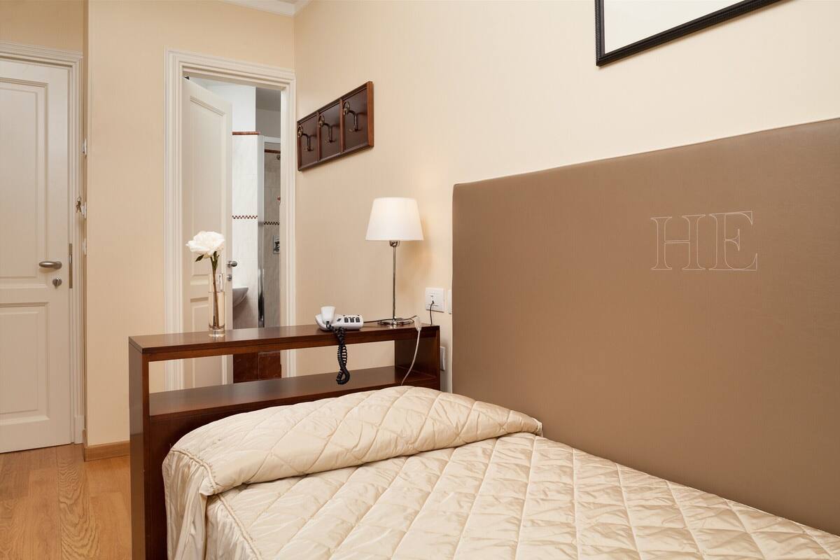A bedroom with a bed, bedside table, lamp, and a wall-mounted phone.