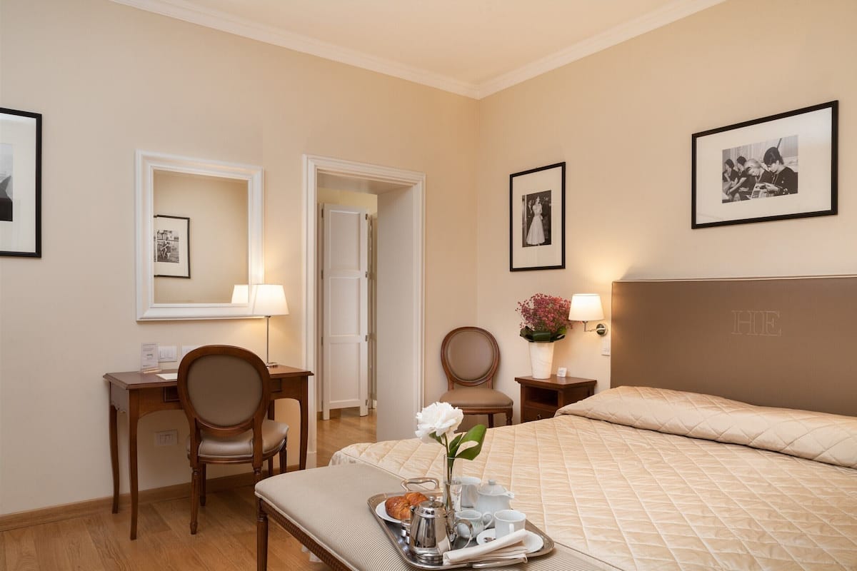 A hotel room with a bed, a desk, a chair, a tray with breakfast items, and framed pictures on the walls.