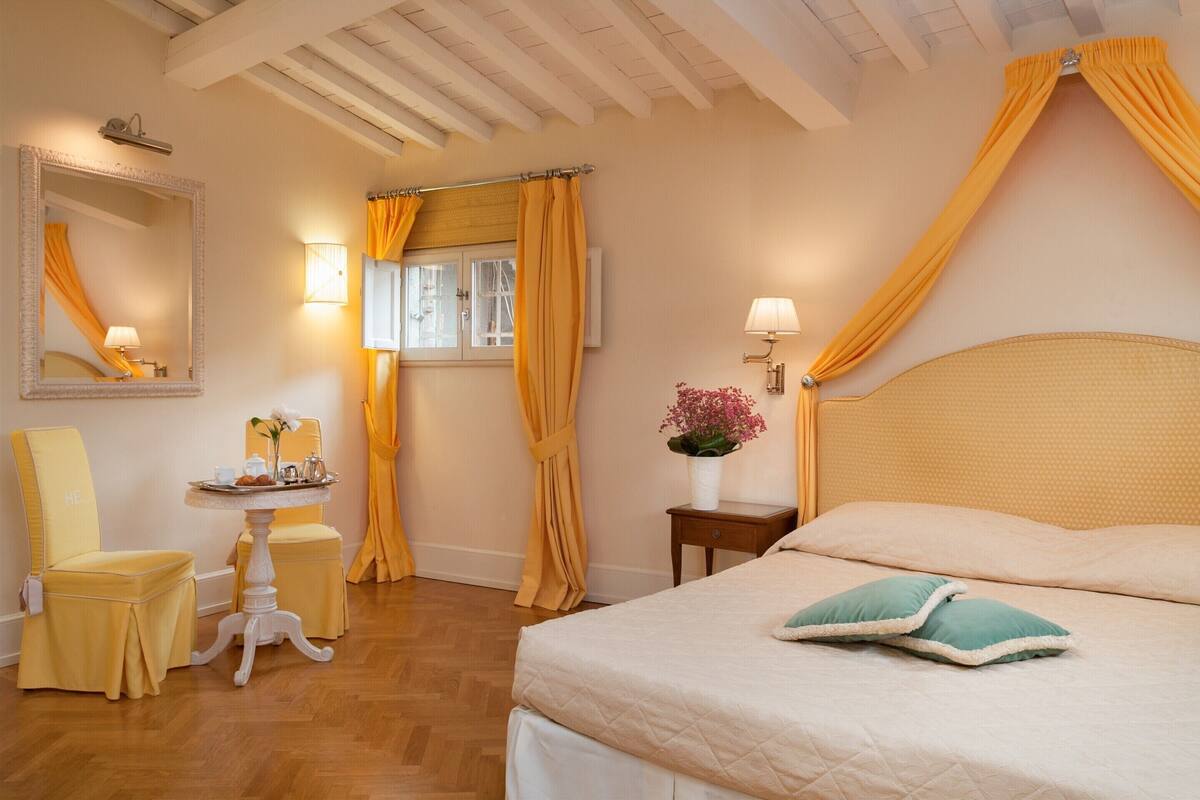 A hotel room with a bed, a chair, a small table with a vase, and a window with curtains.
