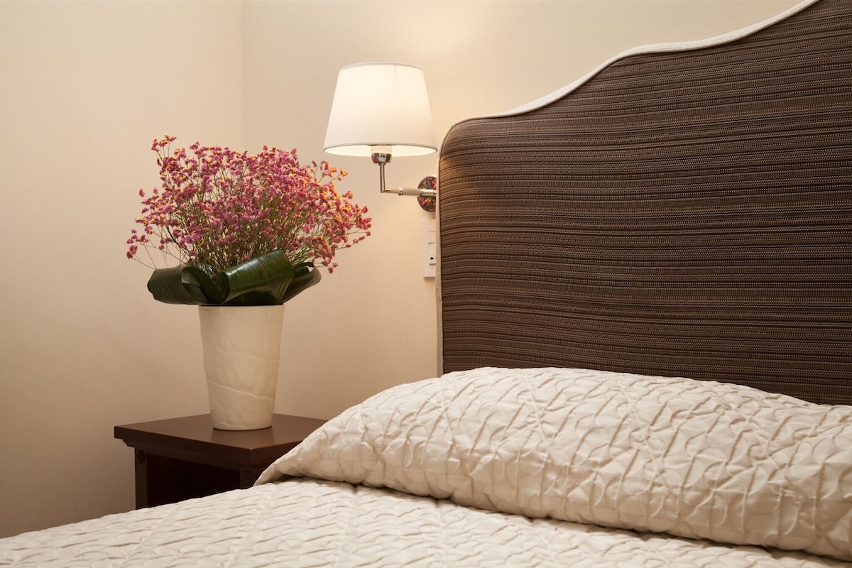 A neatly made bed with a textured white duvet, a bedside table with a white vase of pink flowers, and a wall-mounted lamp.