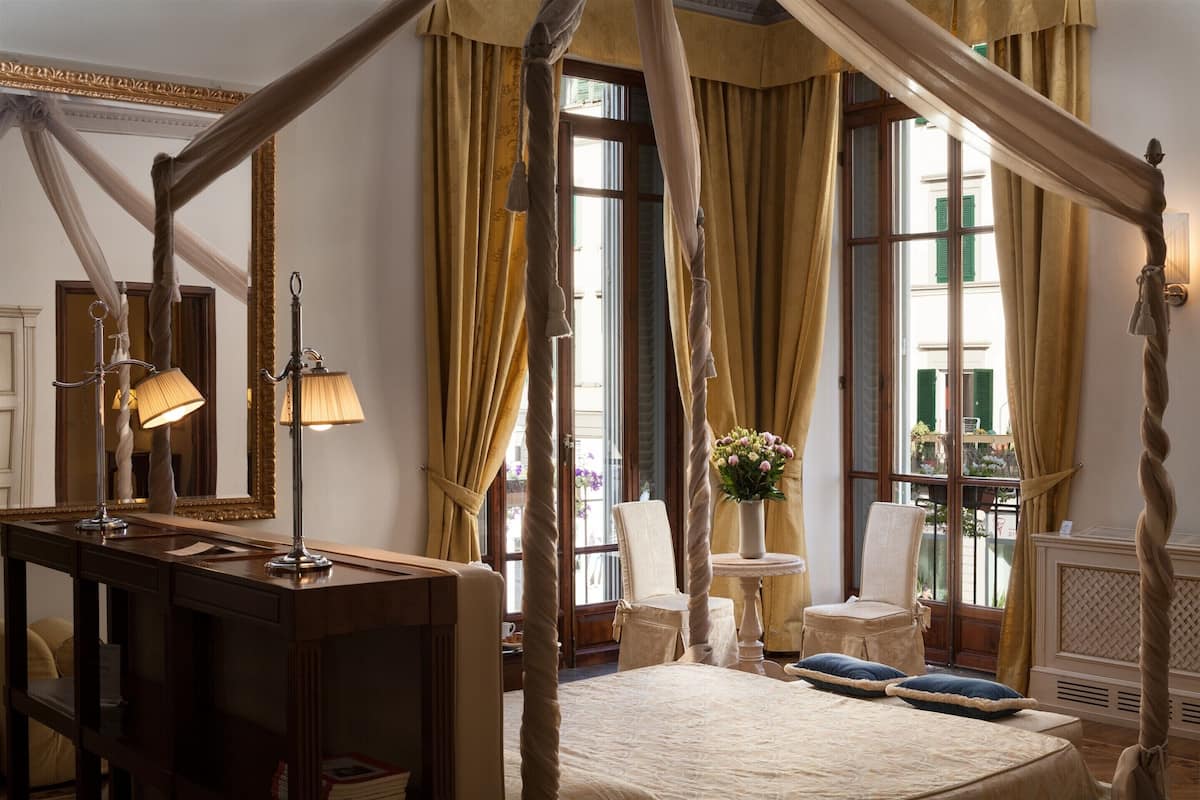 A room with a four-poster bed, a wooden dresser, a table with a vase of flowers, and a chair by the window.