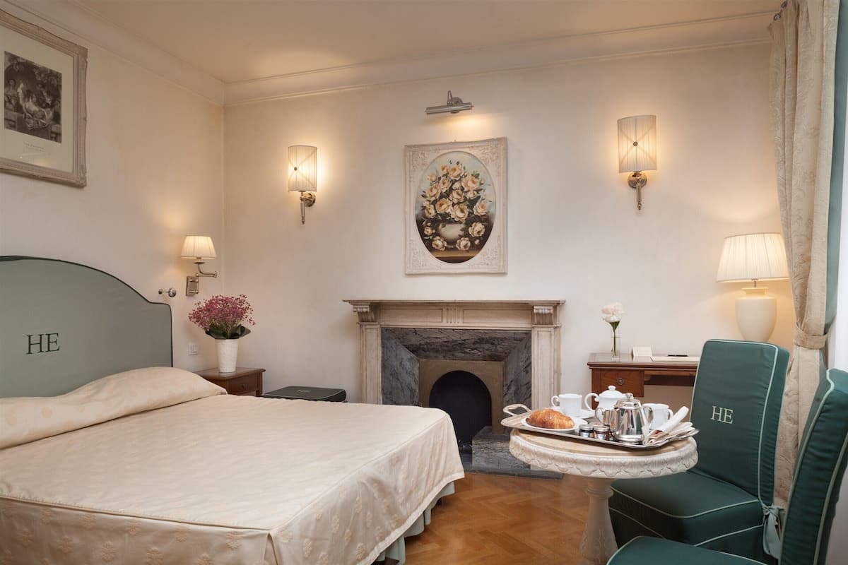 A hotel room with a bed, a fireplace, a dining table with a tray of breakfast, and a chair with "HE" embroidered on it.