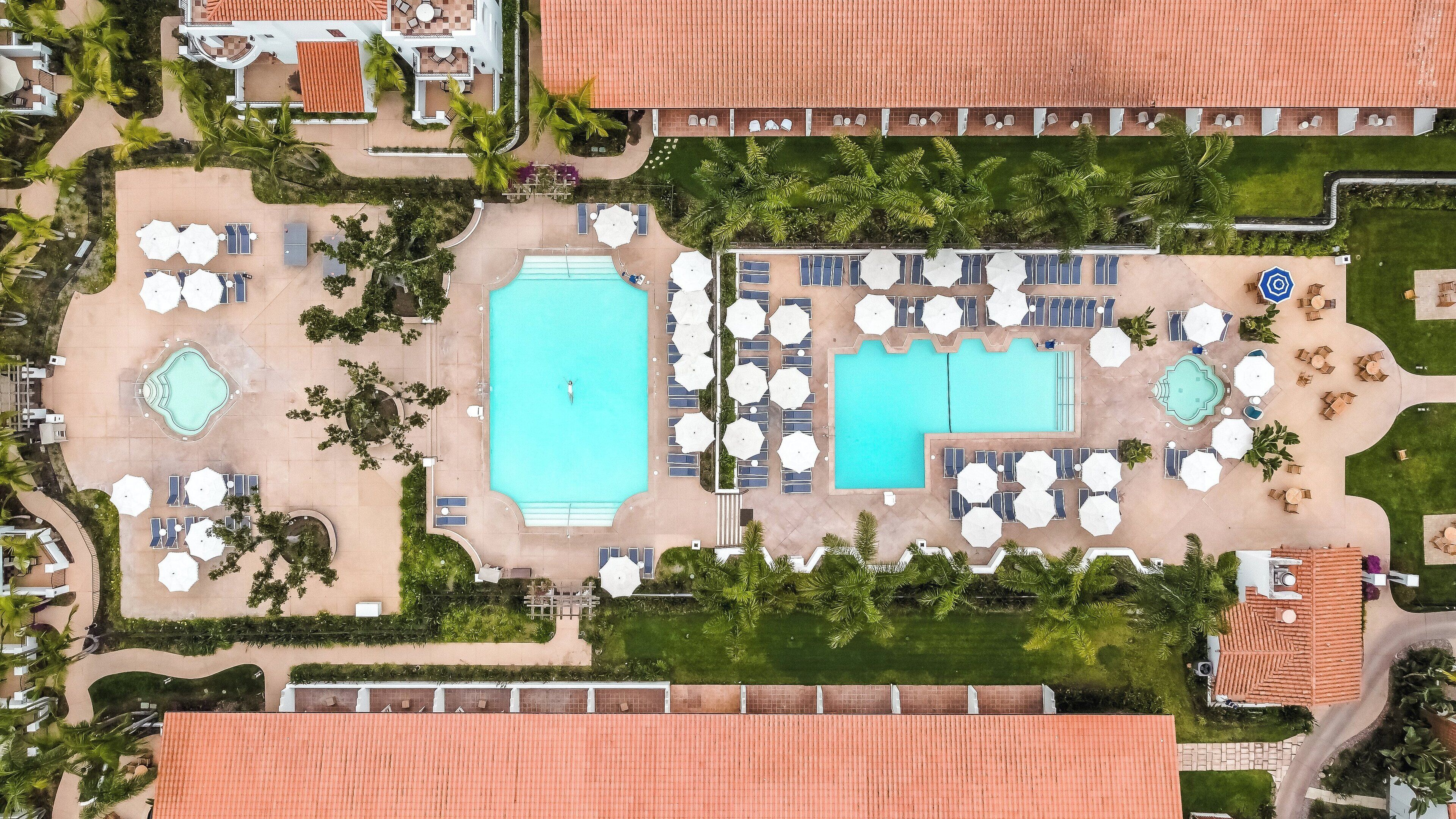 An aerial view of a resort with two swimming pools, sun loungers, and palm trees.