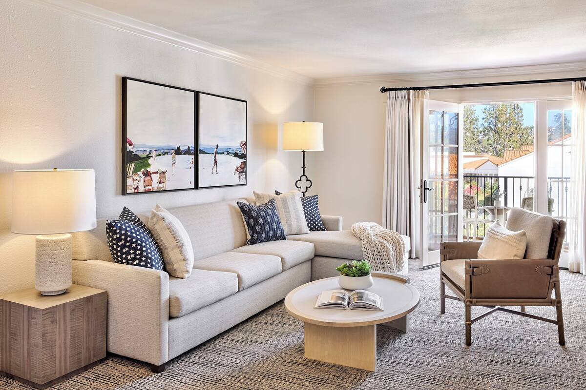 A modern living room with a beige sofa, a round coffee table, and a chair. There is a lamp, a framed picture on the wall, and a view of the outside through a glass door.