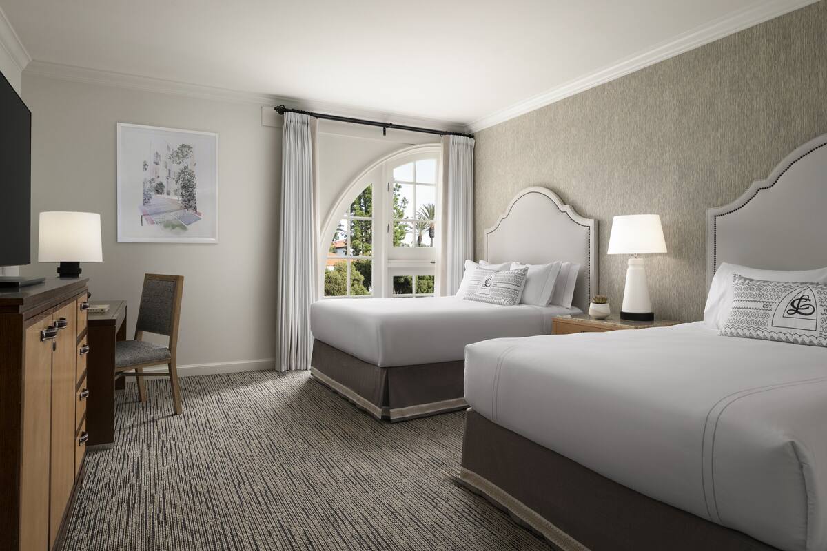 A hotel room with two beds, a desk, and a chair. There is a window with curtains and a view of greenery outside.