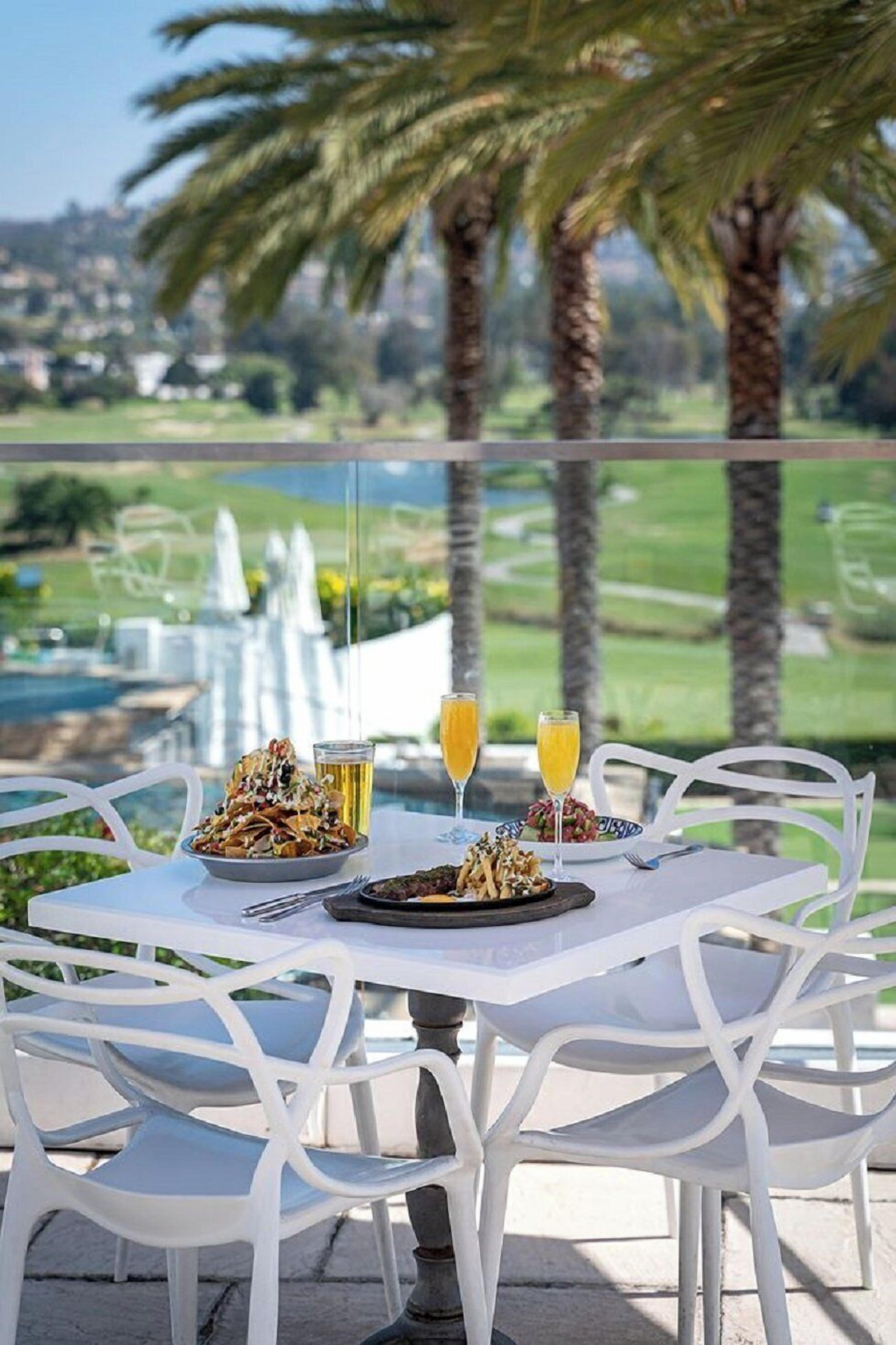 Una mesa de comedor con comida y bebidas, sillas blancas y vistas a un campo de golf y palmeras.
