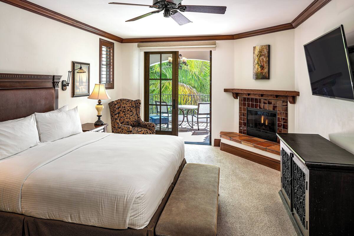 A hotel room with a large bed, a ceiling fan, a fireplace, a TV, and a view of the outdoors.