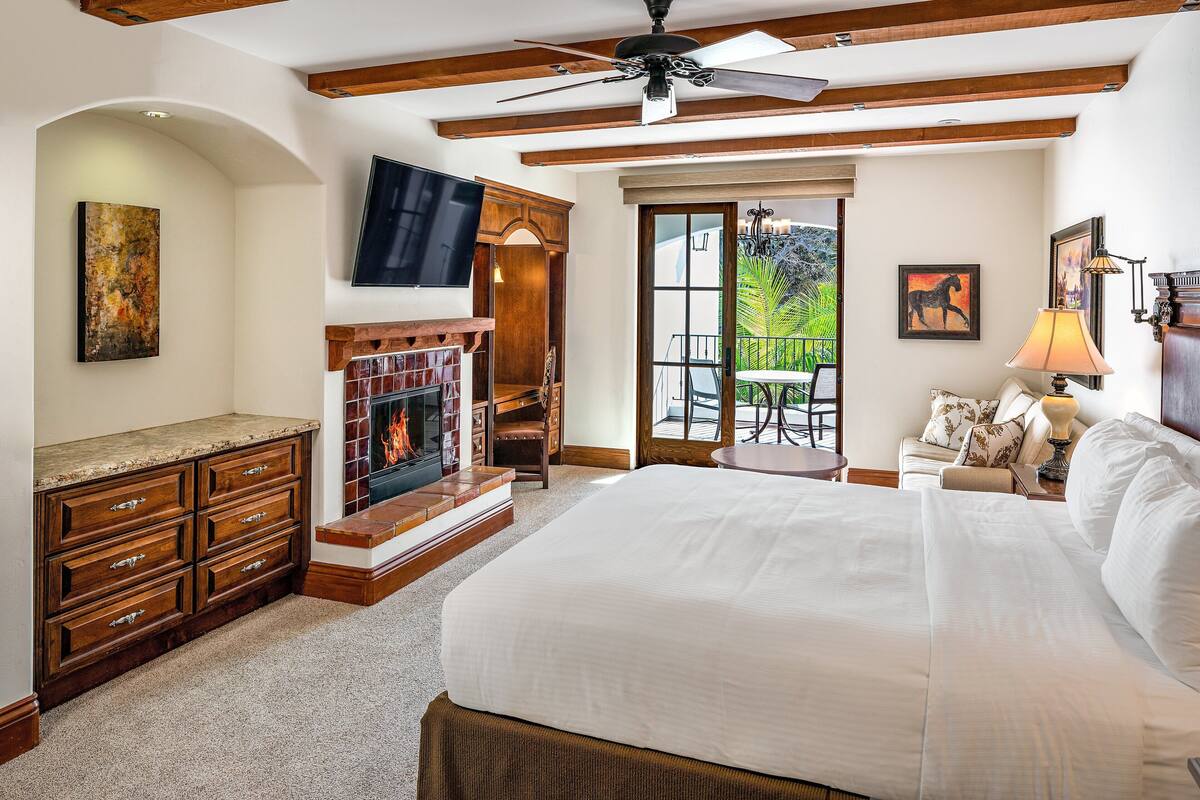 A spacious bedroom with a large bed, a fireplace, a TV, and a balcony with outdoor seating.