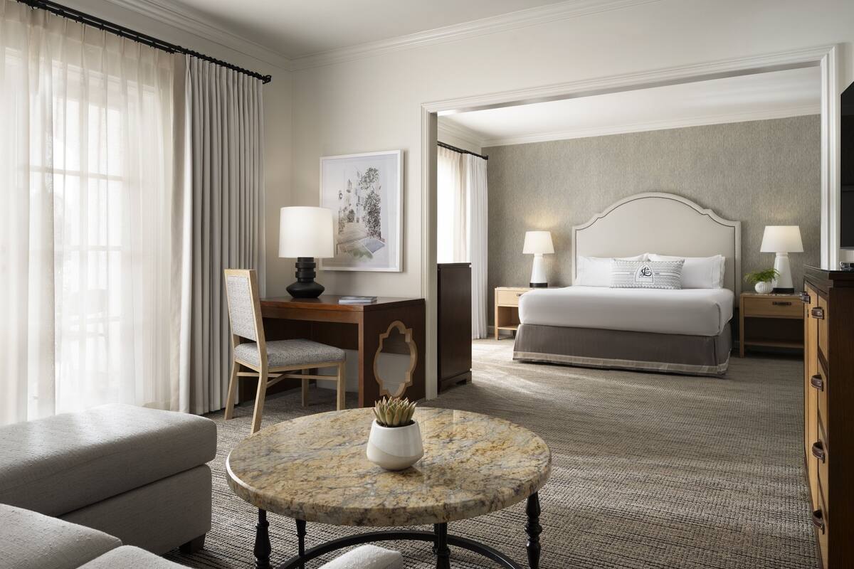 A hotel room with a bed, a desk with a chair, a round table with a plant, and a large window with curtains.