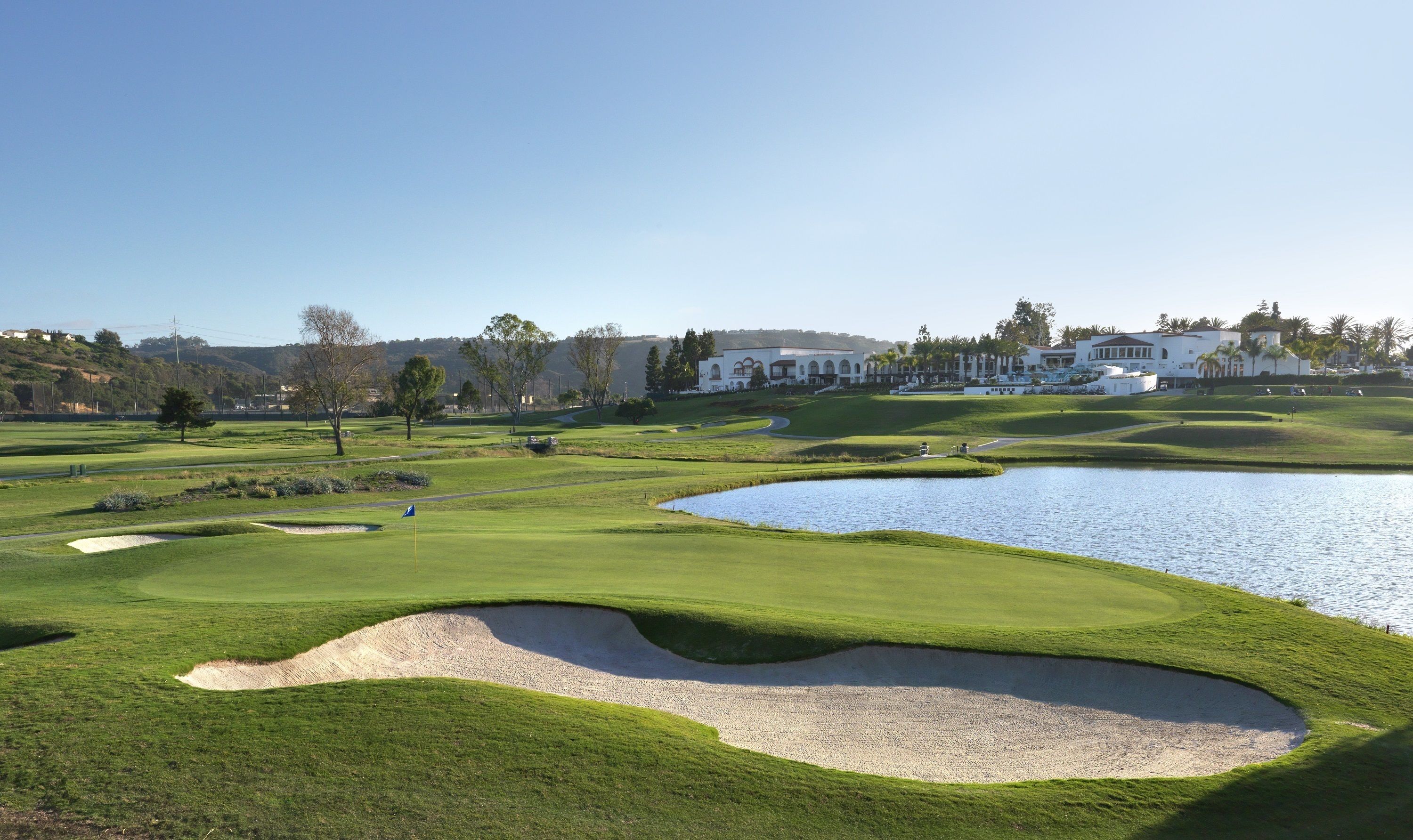 Um campo de golfe com bunkers, lago e um clube de campo ao fundo.