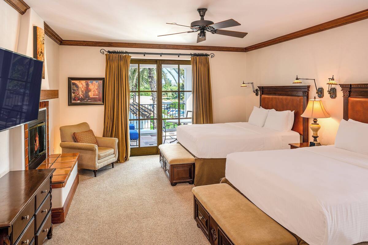 A hotel room with two beds, a TV, a chair, a bench, and a balcony with curtains.