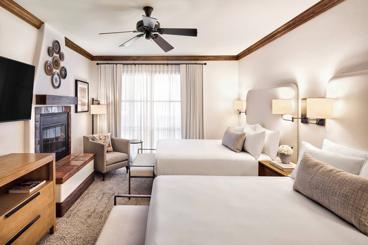 A hotel room with a bed, a chair, a desk, a TV, a ceiling fan, and a window with curtains.