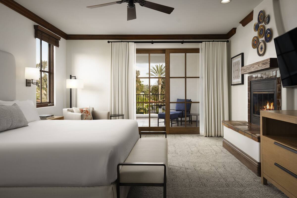 A hotel room with a large bed, a bench, a fireplace, a TV, and a balcony with a view of palm trees.
