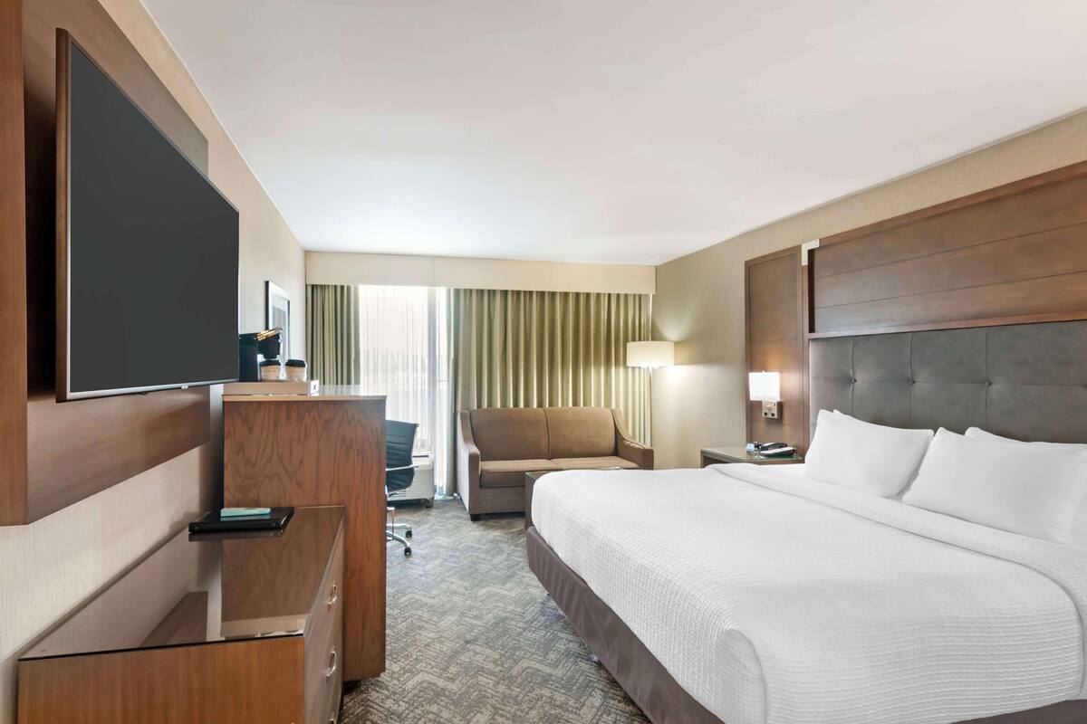 A hotel room with a large bed, a desk with a flat-screen TV, a chair, a lamp, and a view of the outdoors through a window with curtains.