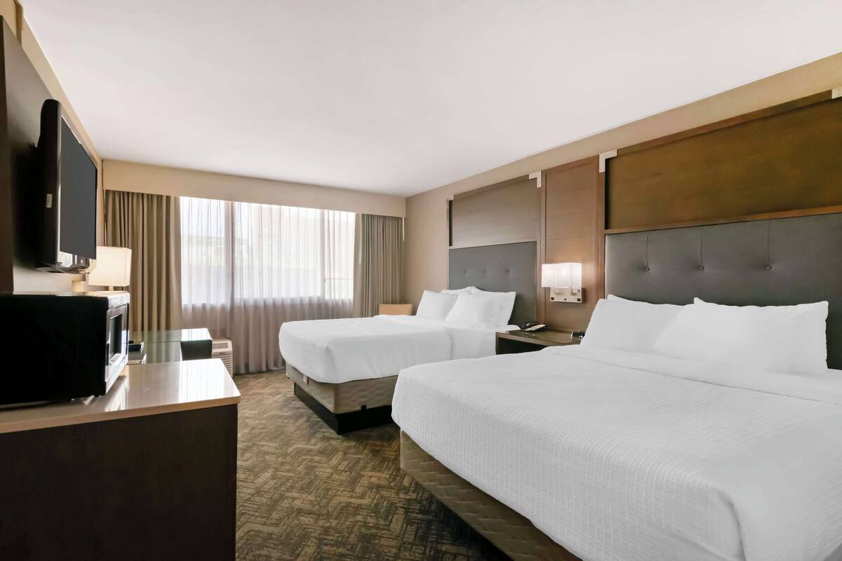 A hotel room with two beds, a desk with a flat-screen TV, a minibar, and a large window with curtains.