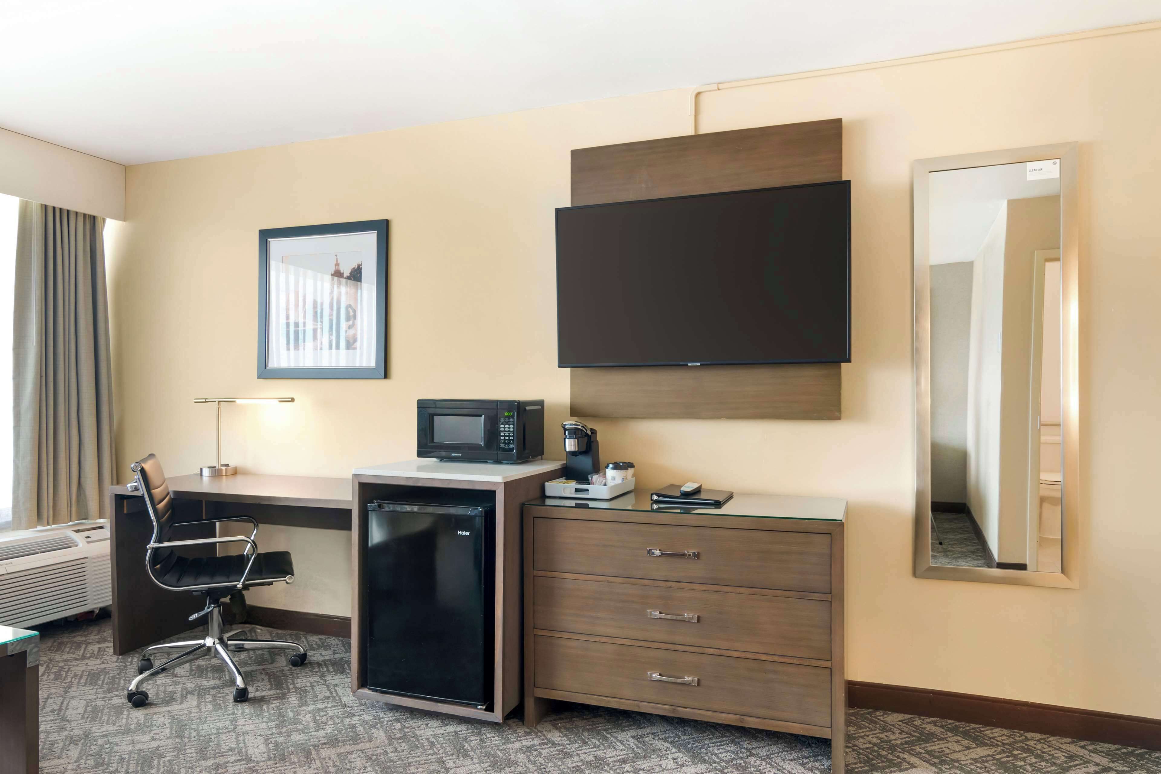 A hotel room with a desk, chair, microwave, coffee maker, refrigerator, and a large mirror.