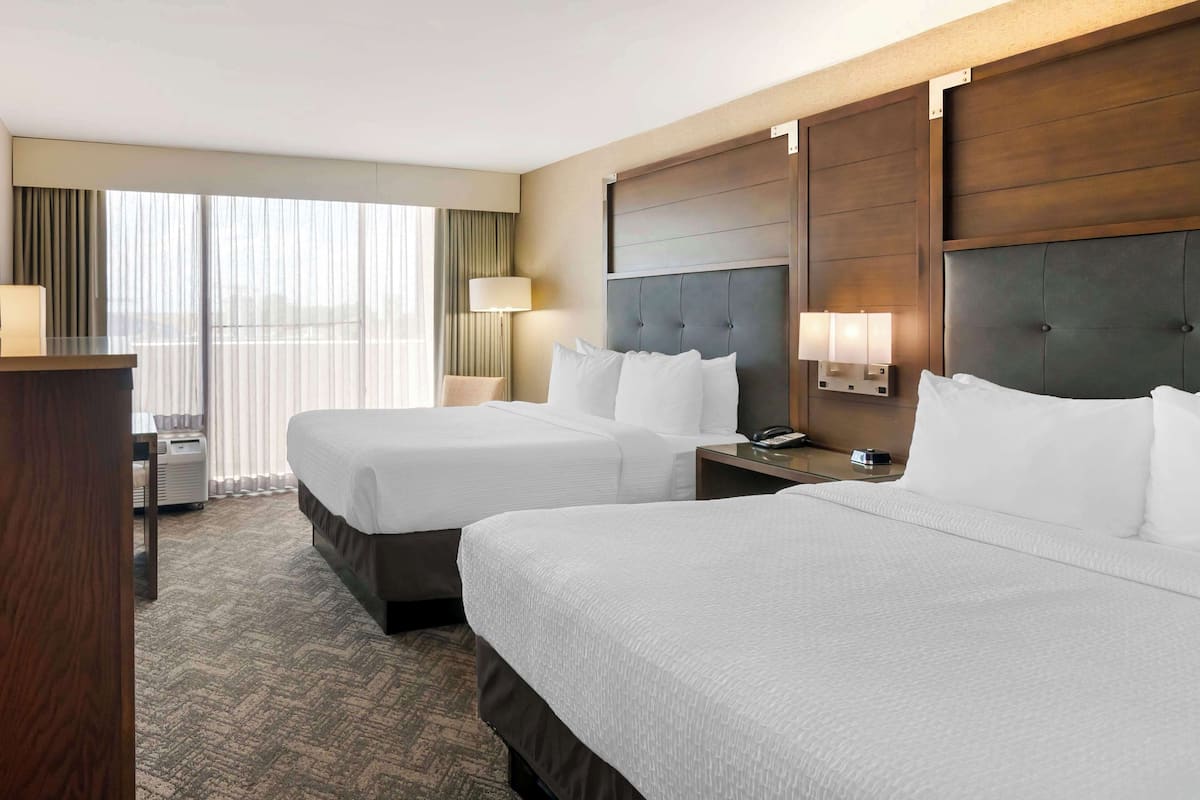 A hotel room with two beds, a desk, a lamp, and a view of the city through large windows.