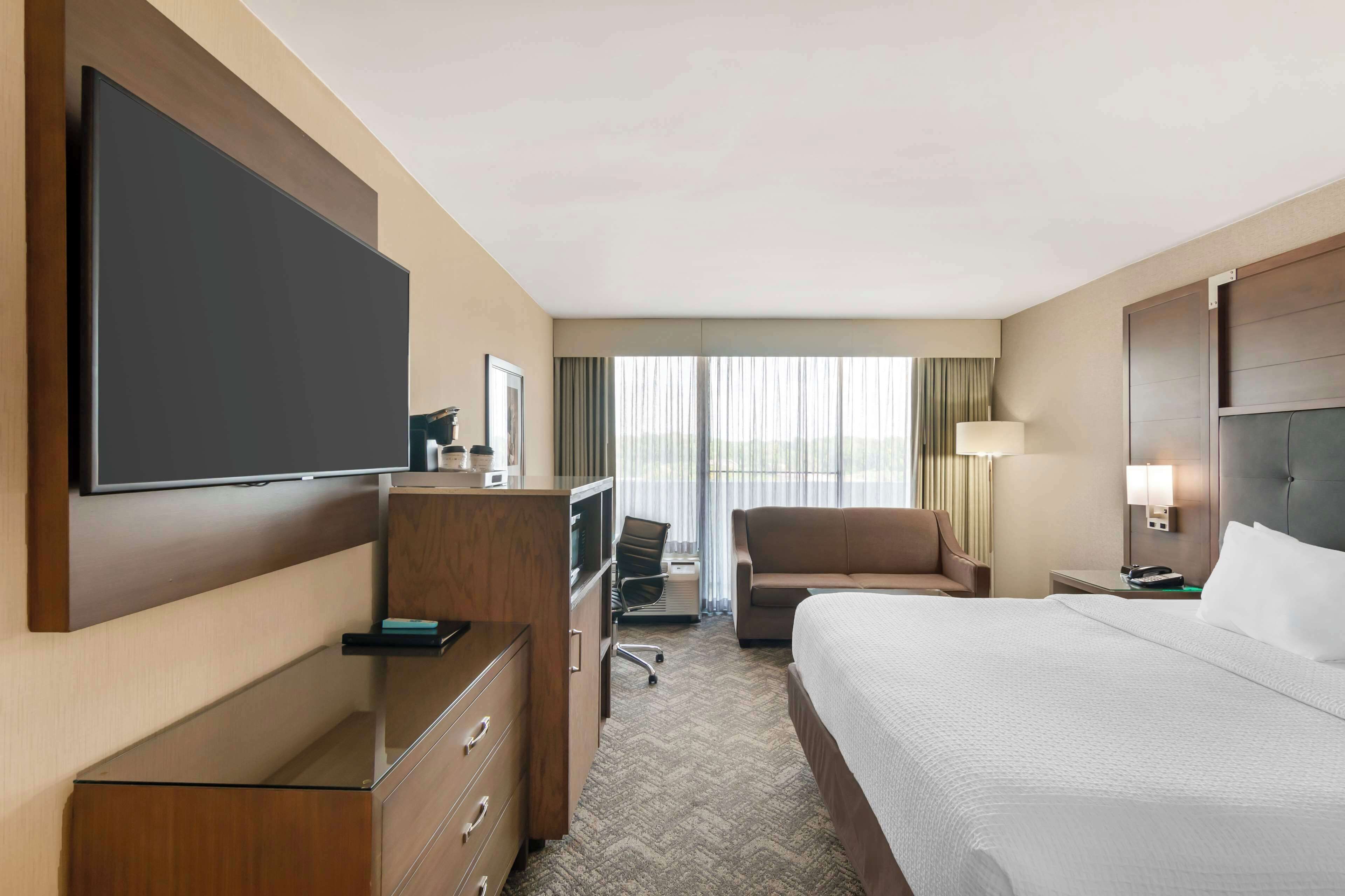 A hotel room with a large bed, a desk with a flat-screen TV, a chair, a desk lamp, and a window with curtains.