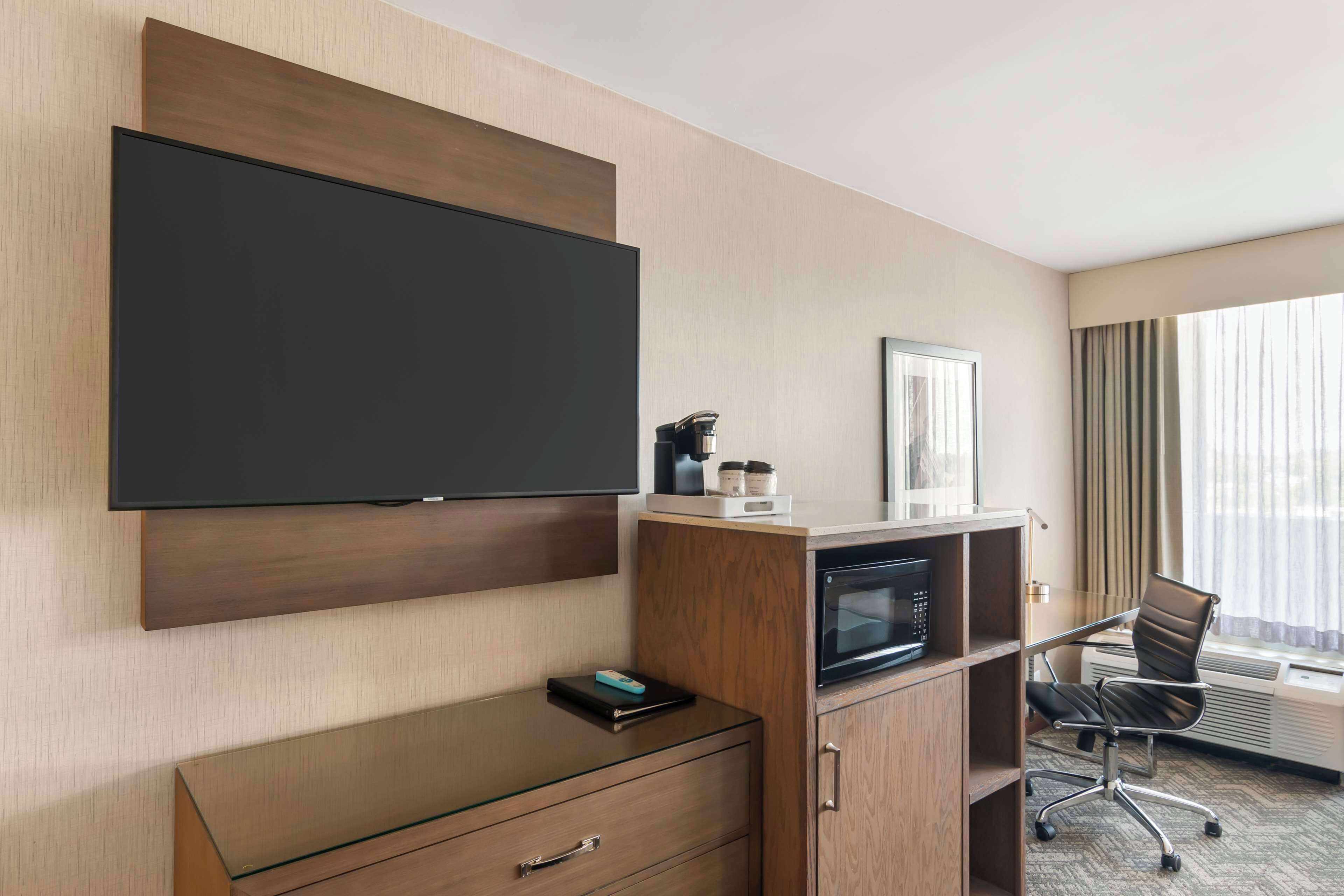 Une chambre d’hôtel équipée d’un téléviseur à écran plat fixé au mur, d’un bureau en bois avec un ordinateur portable, d’un four à micro-ondes et d’une chaise.