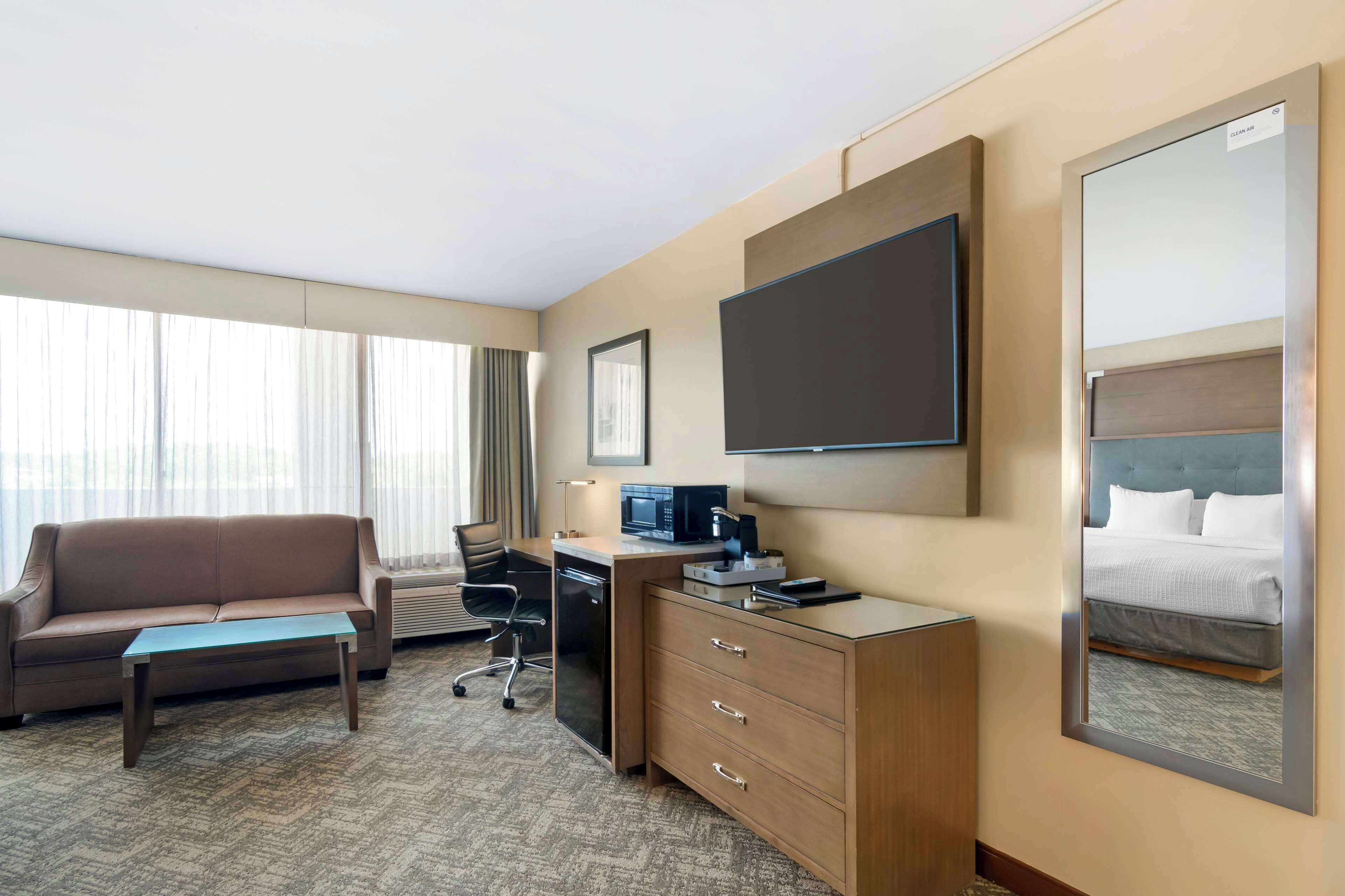 A hotel room with a sofa, a desk with a computer, a flat-screen TV, a dresser, and a large mirror.