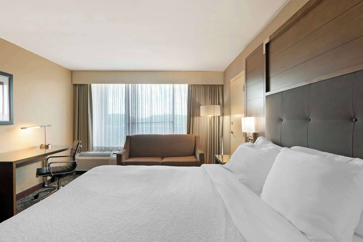 A hotel room with a large bed, a desk with a chair, a sofa, and a window with curtains.