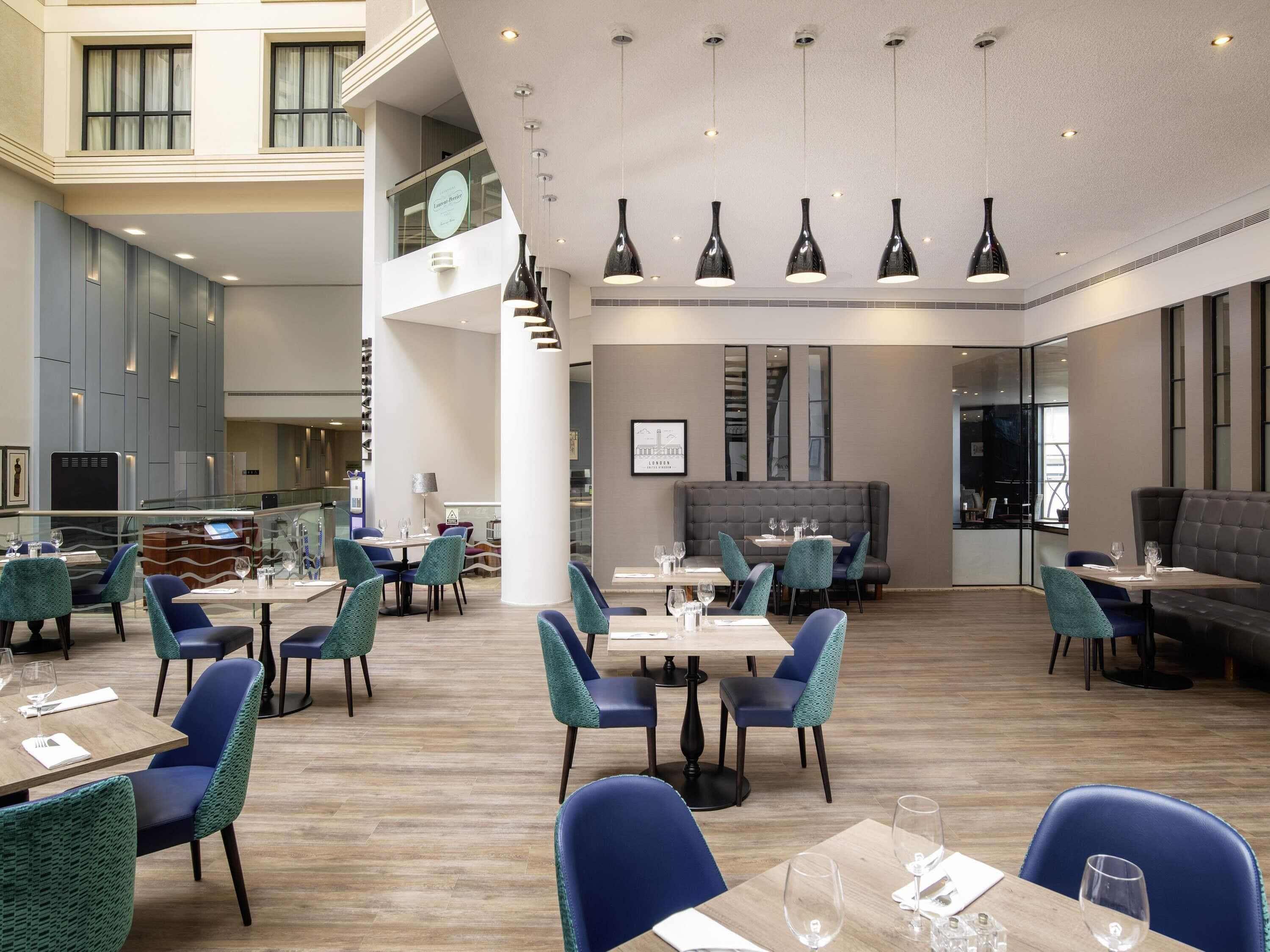 En modern hotellobby med flera matplatser, inklusive en bar och en sittgrupp med högt i tak och hängande lampor.