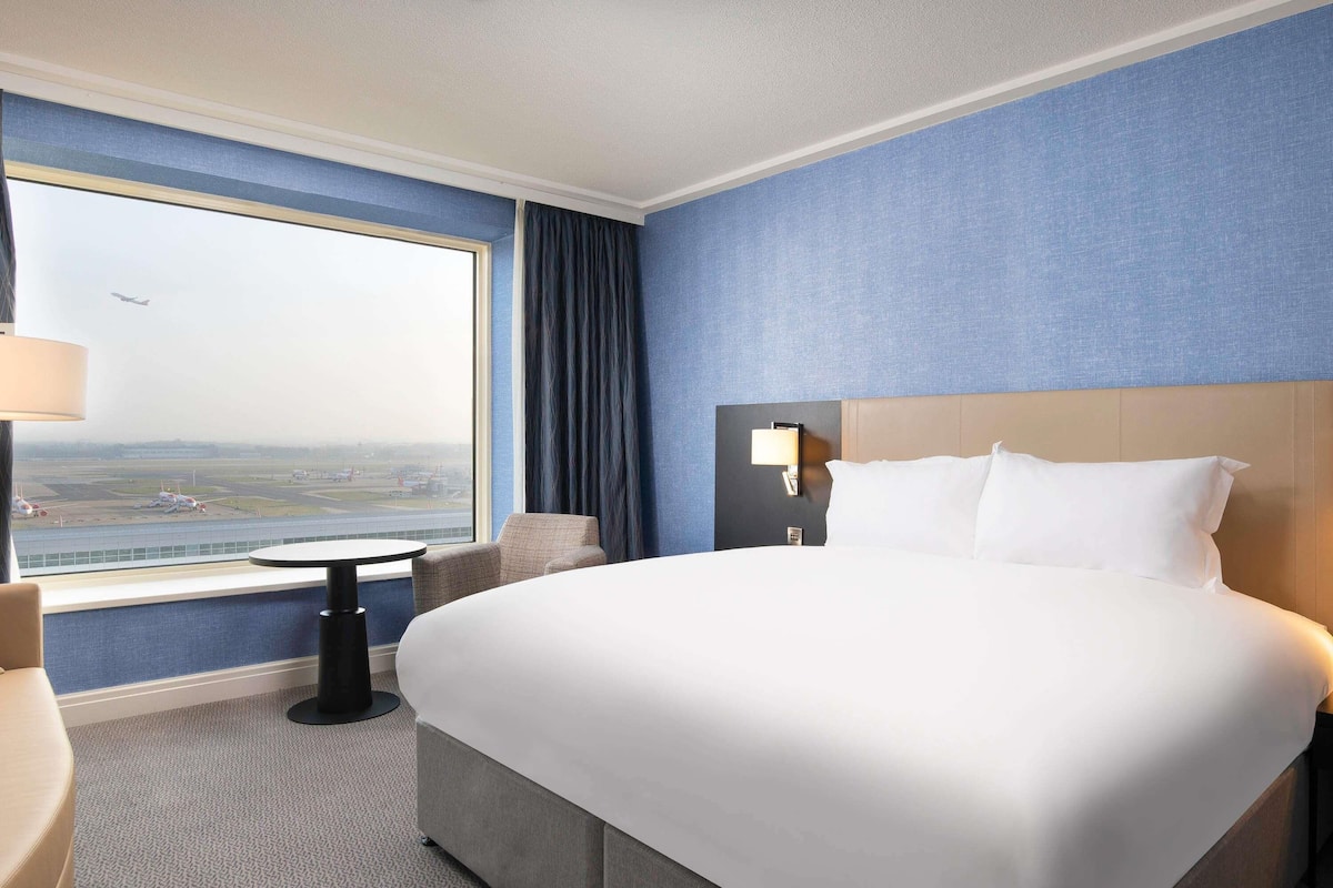 A hotel room with a large bed, a view of an airport, a small table, and a chair.