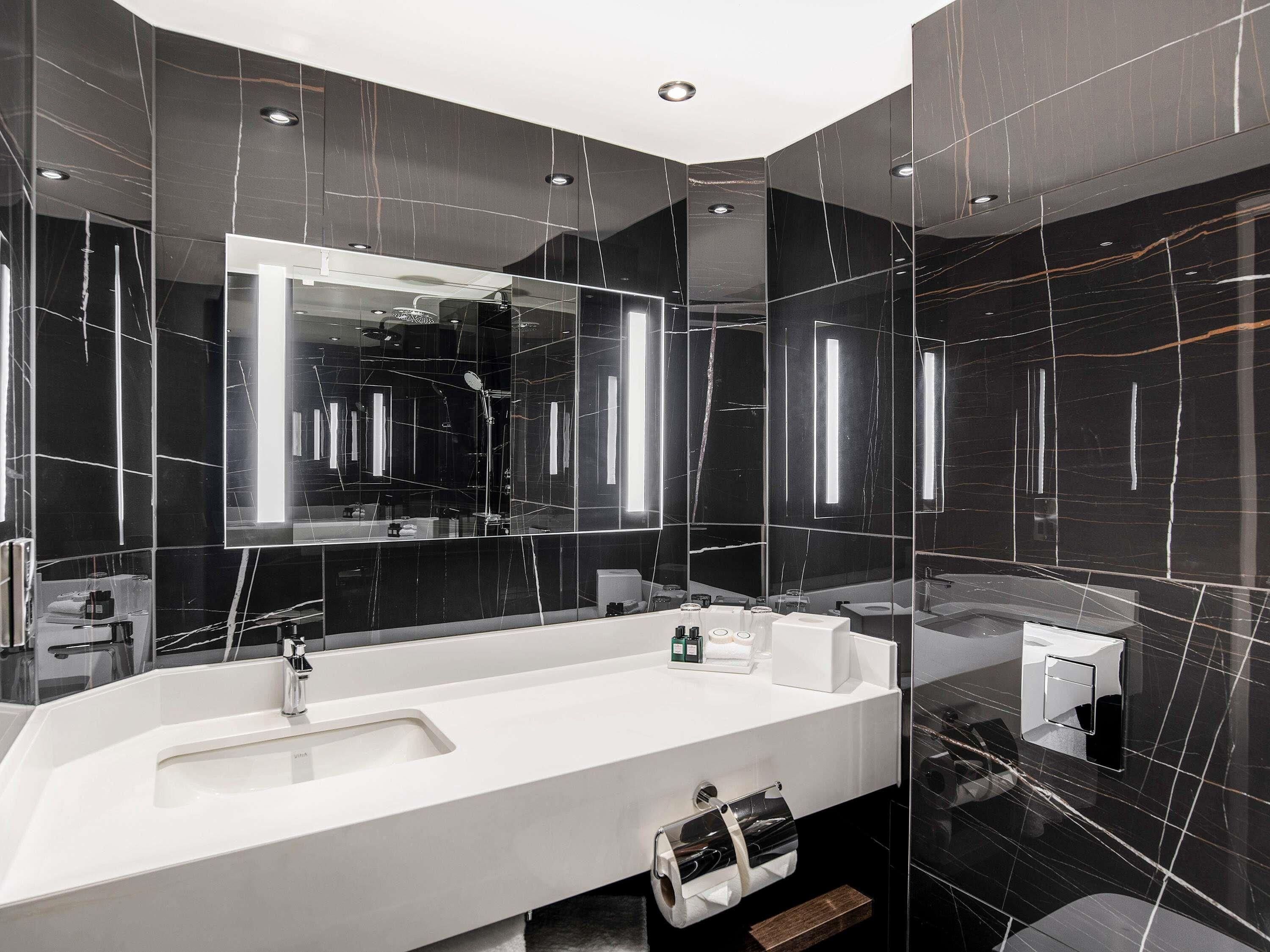 Un baño moderno con un espejo grande, lavamanos blanco y paredes de mármol negro.