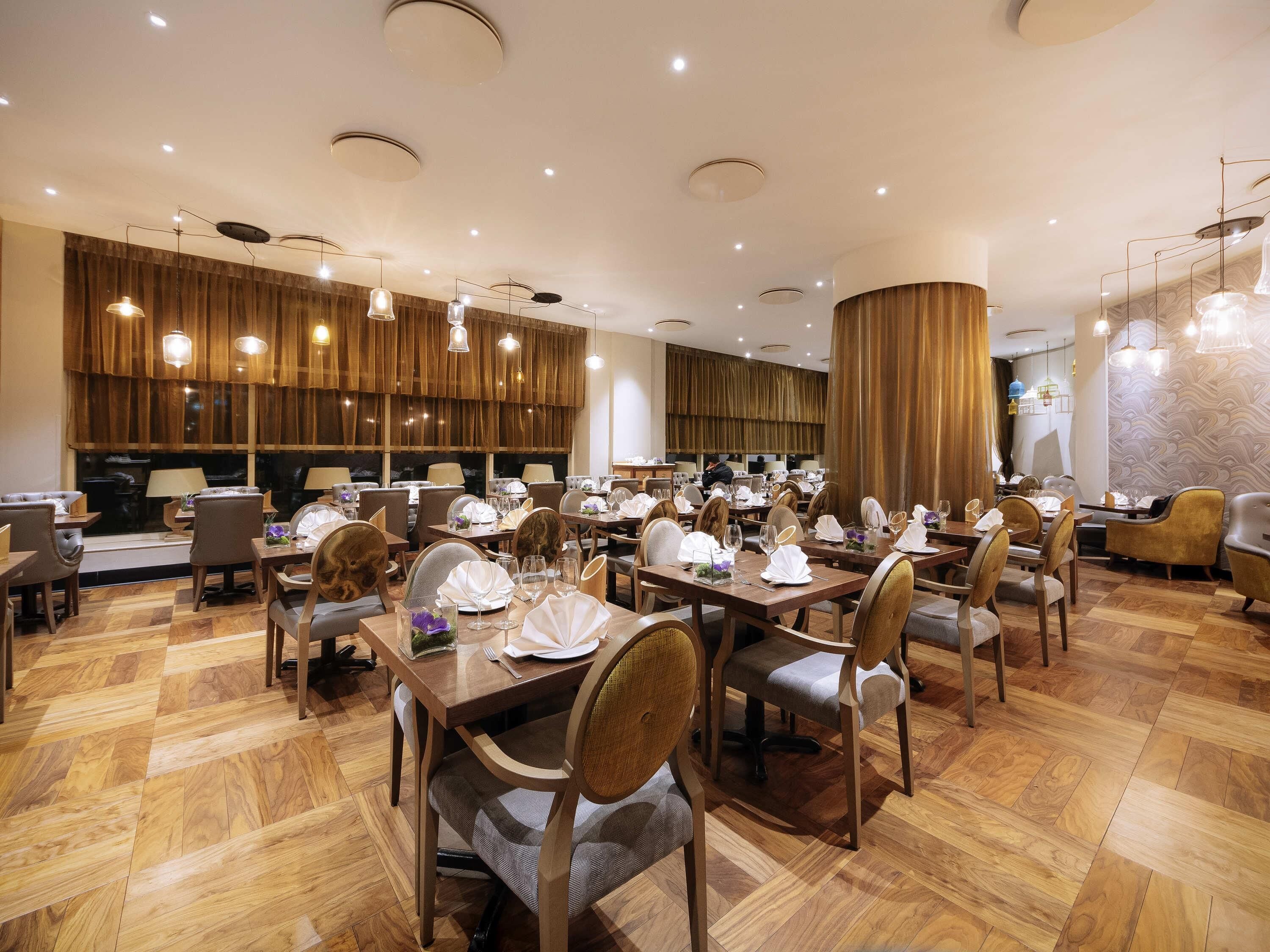 Comedor de hotel con varias mesas y sillas, luces en el techo y un ventanal con cortinas.