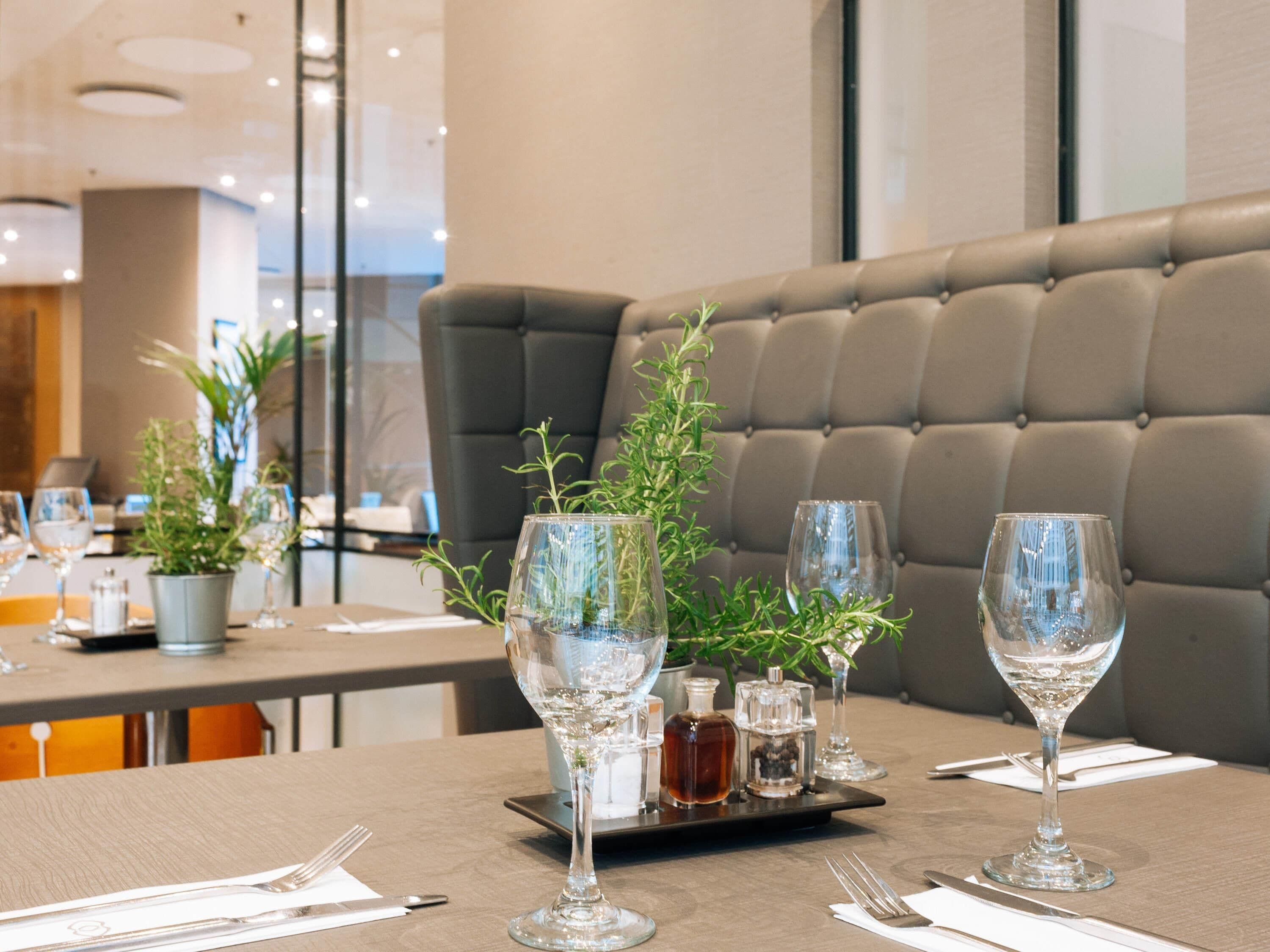 Un comedor con mesas dispuestas para dos, con cristalería transparente, una bandeja con salsas y una planta en maceta.