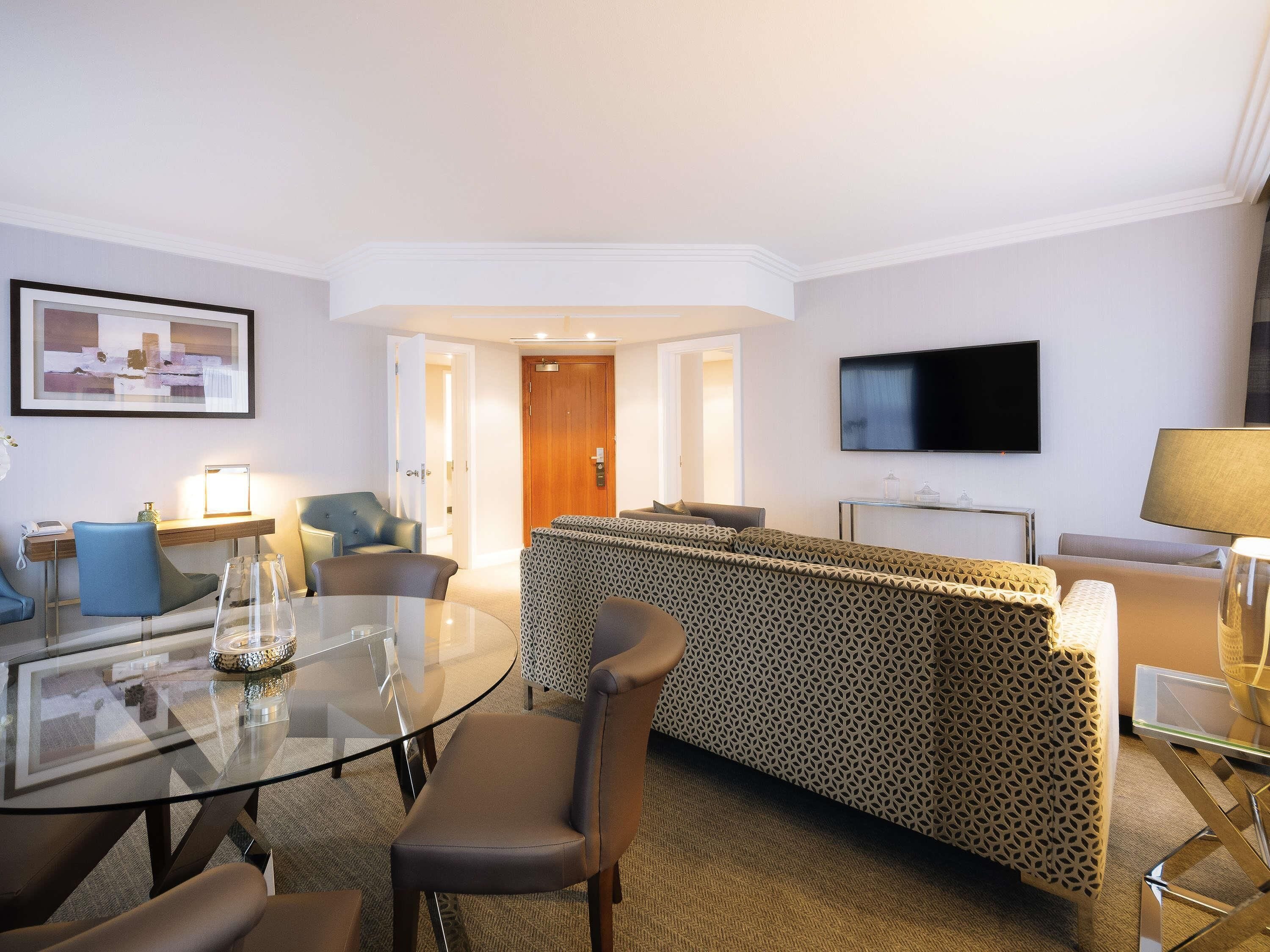 A modern hotel room with a glass dining table, a sofa, a TV, and a door leading to another room.