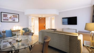 A modern hotel room with a glass dining table, a sofa, a TV, and a door leading to another room.