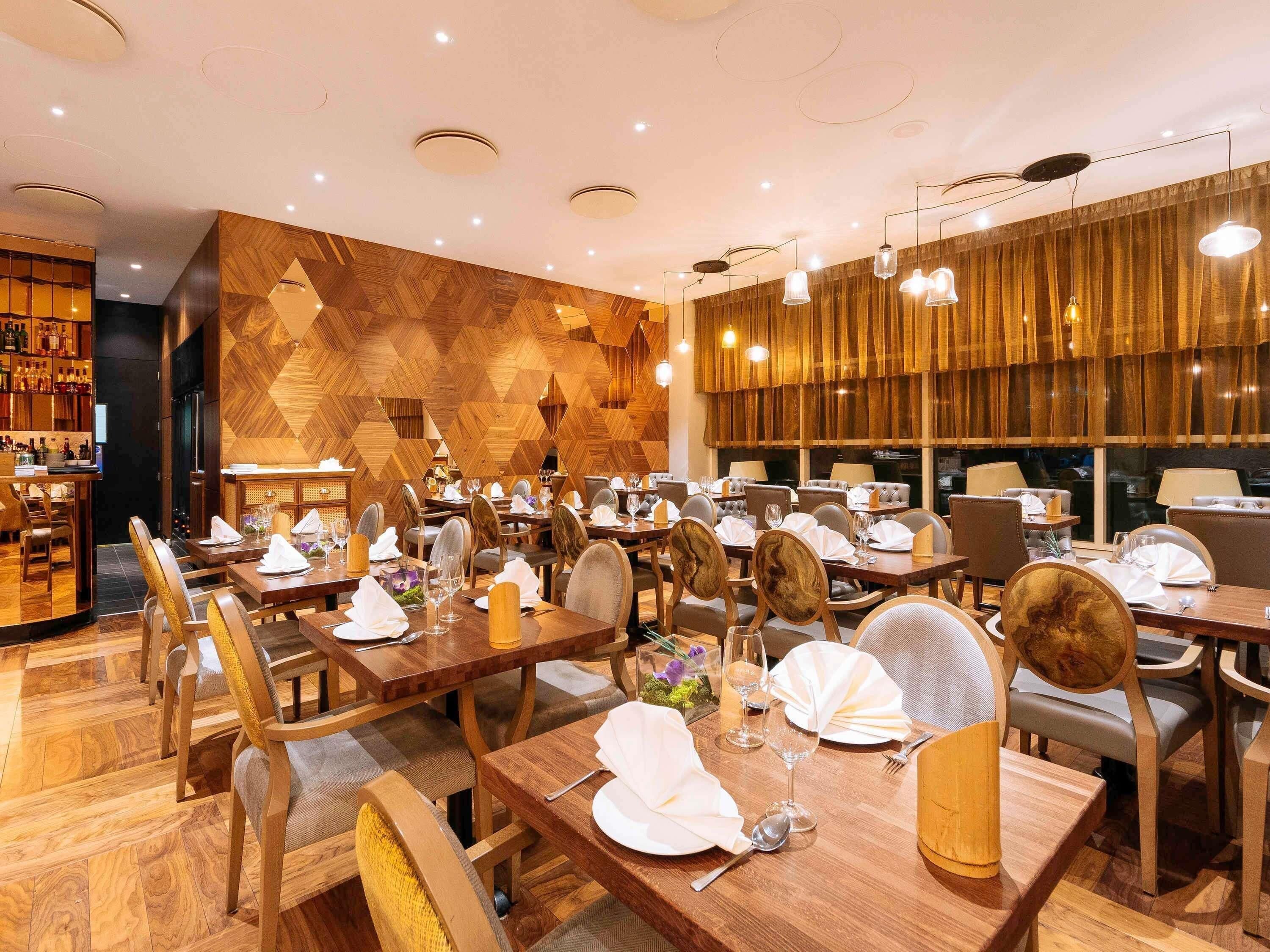 A restaurant with wooden flooring, tables set with white tablecloths, and a bar area with shelves of bottles.