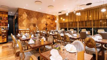 A restaurant with wooden flooring, tables set with white tablecloths, and a bar area with shelves of bottles.