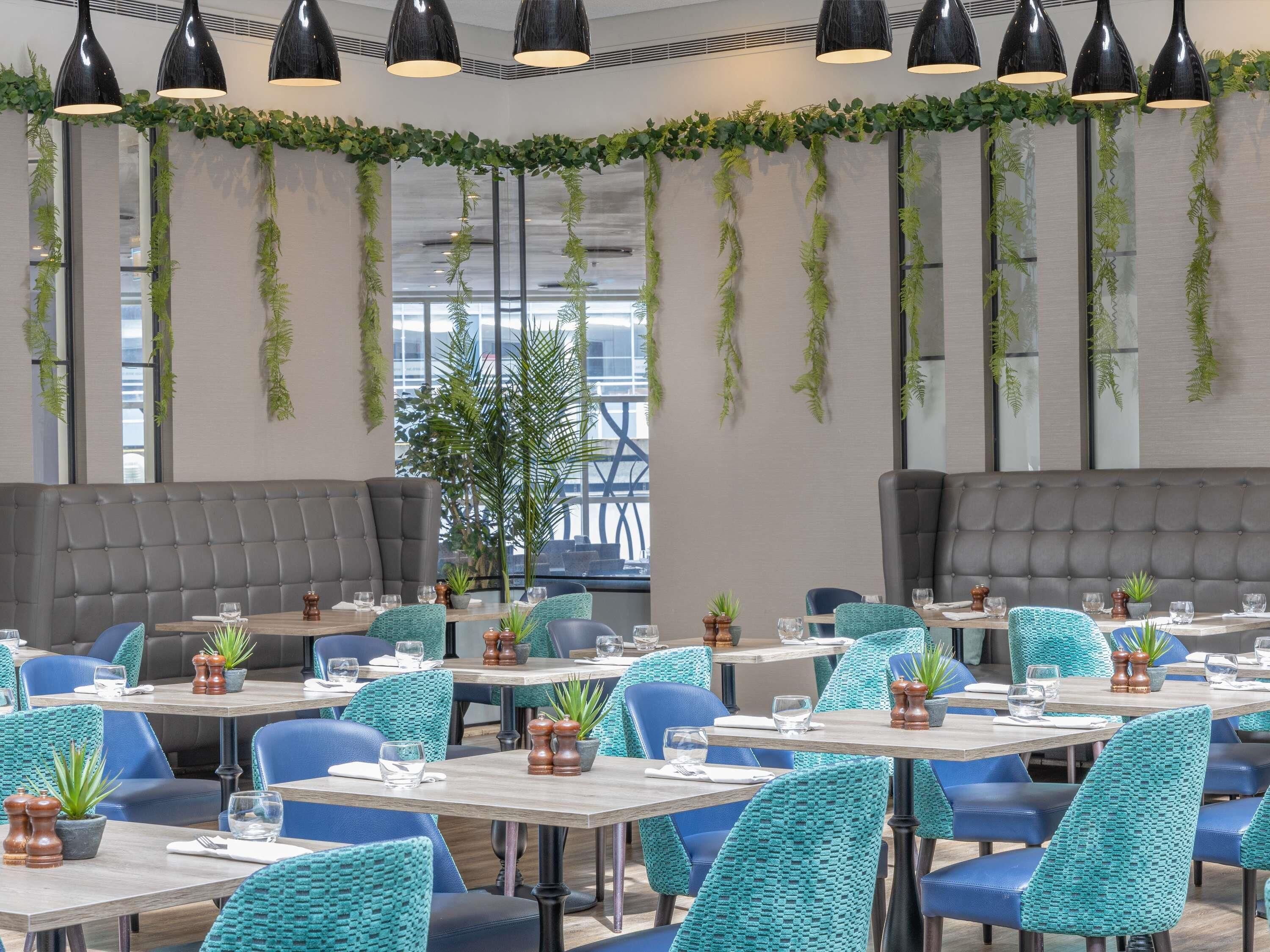 Un interior de restaurante moderno con mesas altas, sillas tapizadas en azul y plantas verdes.