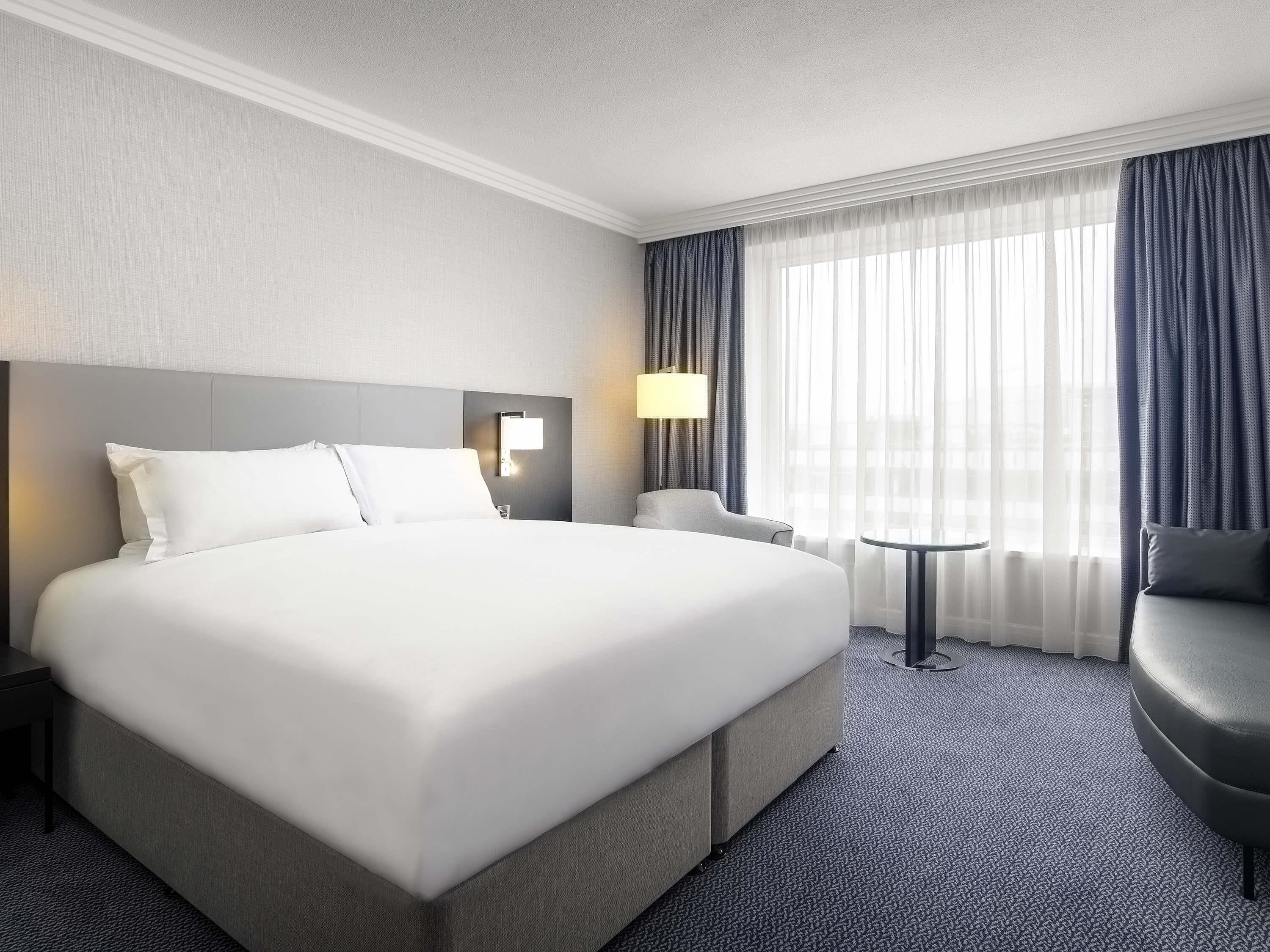 A hotel room with a large bed, a desk, a chair, and a view of the city through sheer curtains.