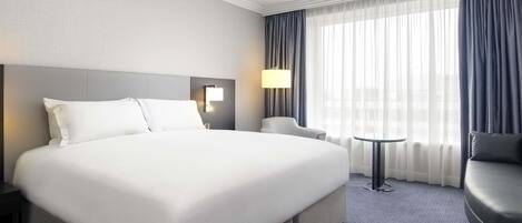 A hotel room with a large bed, a desk, a chair, and a view of the city through sheer curtains.