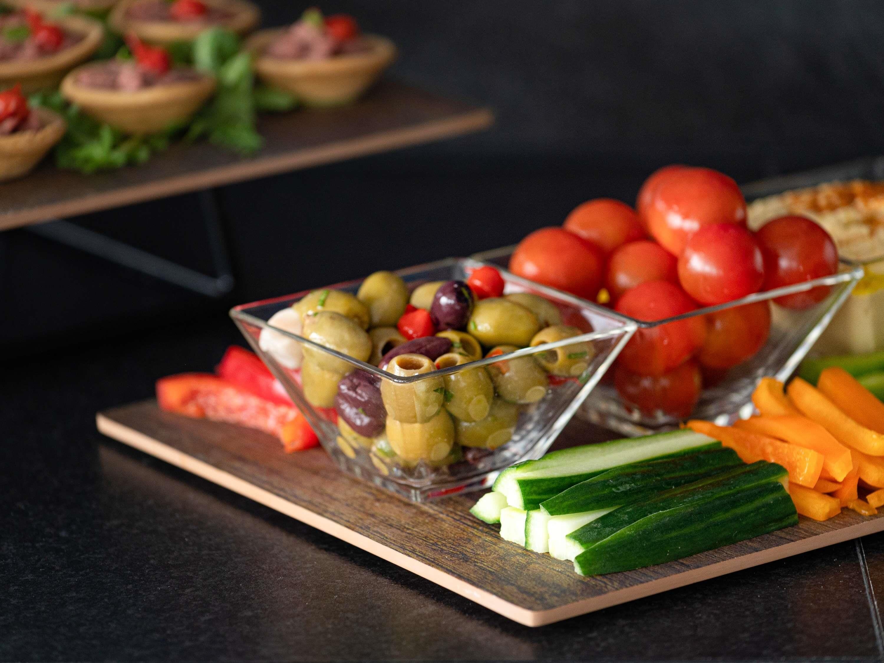 A glass bowl filled with mixed olives and cherry tomatoes on a wooden board, with sliced cucumbers and carrots beside it.
