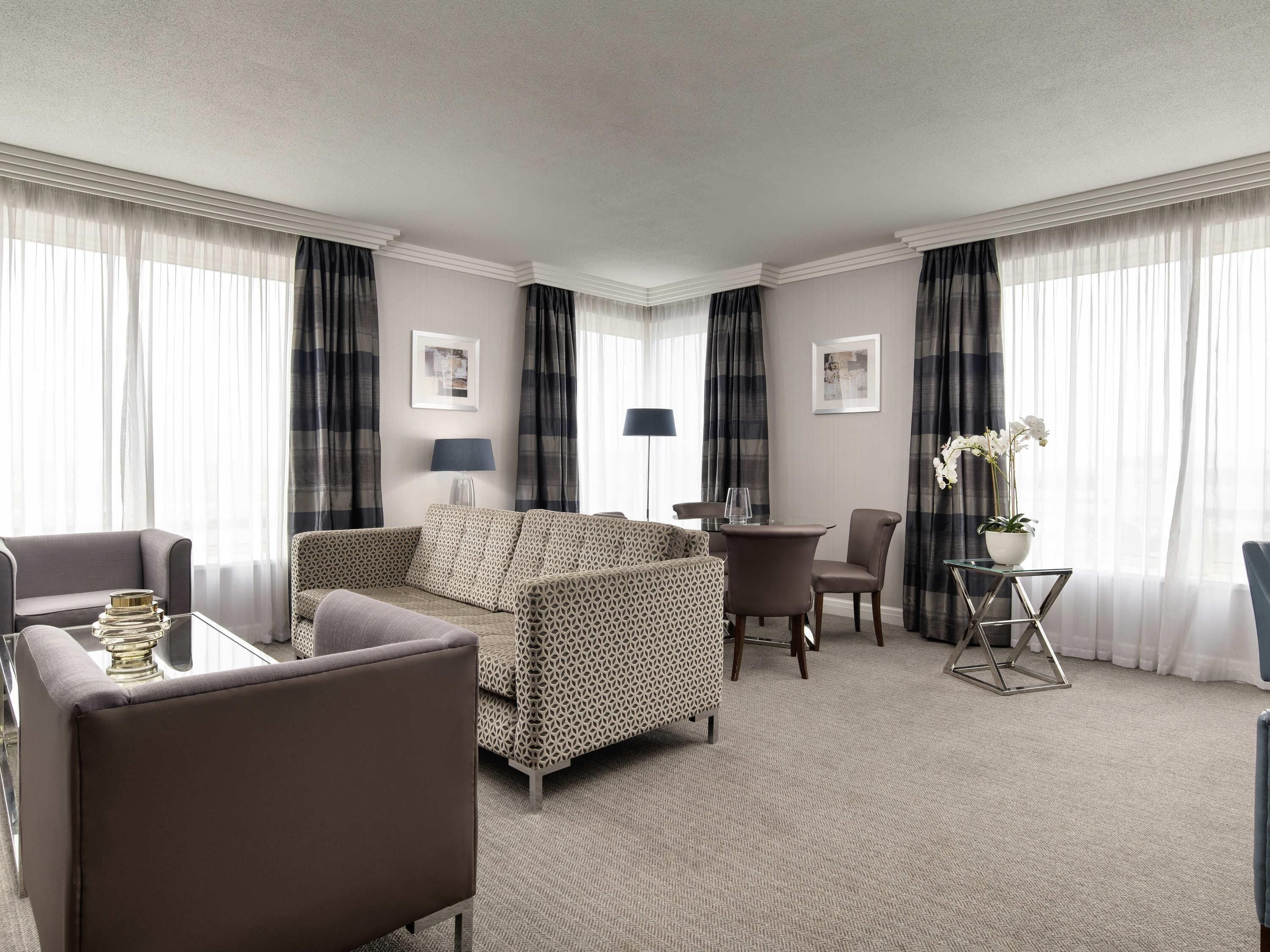 A hotel room with a sofa, armchairs, a small table, a lamp, and a potted plant.