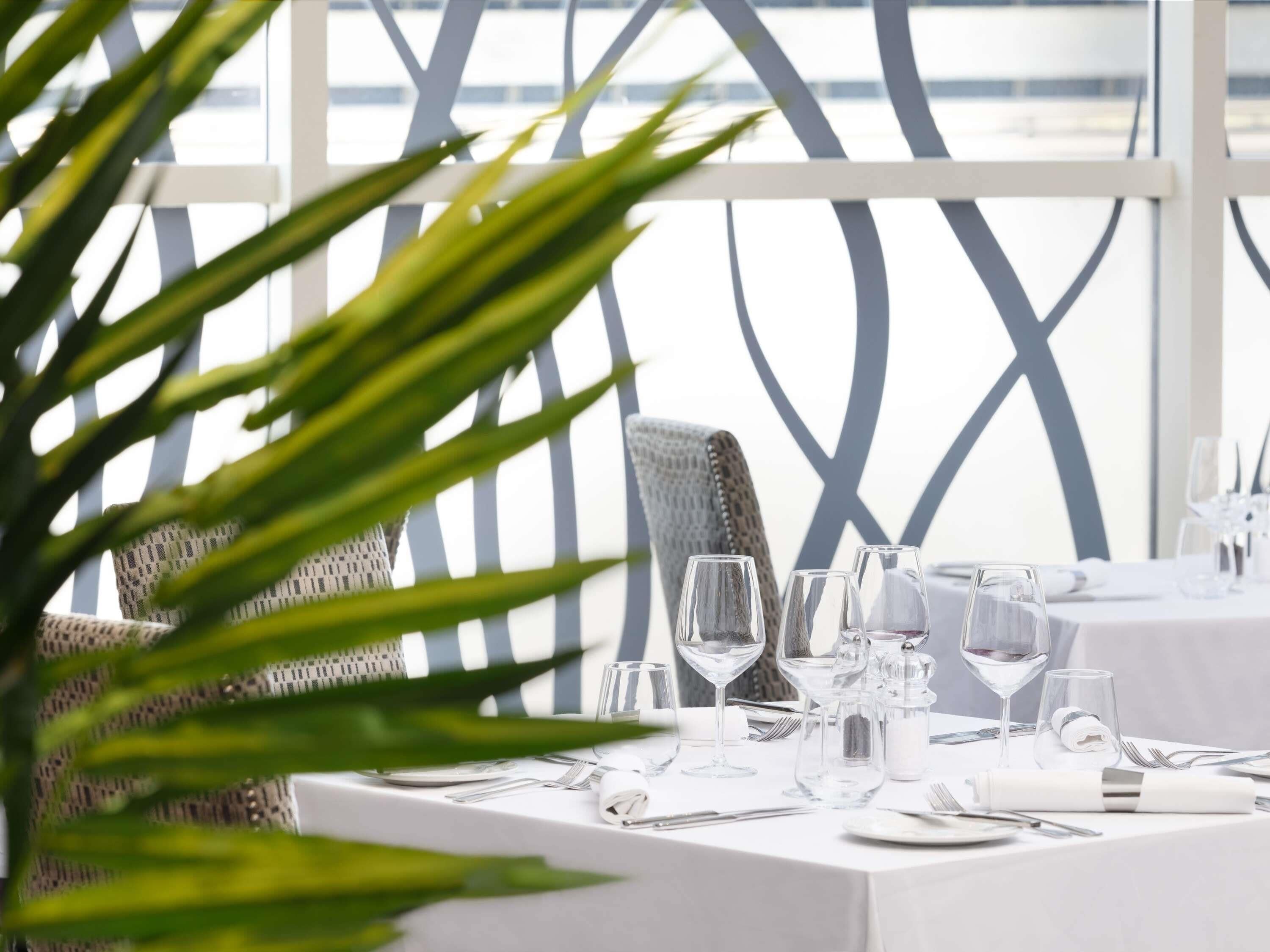 Mesa lista para comer con cristalería transparente y cubiertos, una silla con estampado y vista a través de una estructura de ventana geométrica.
