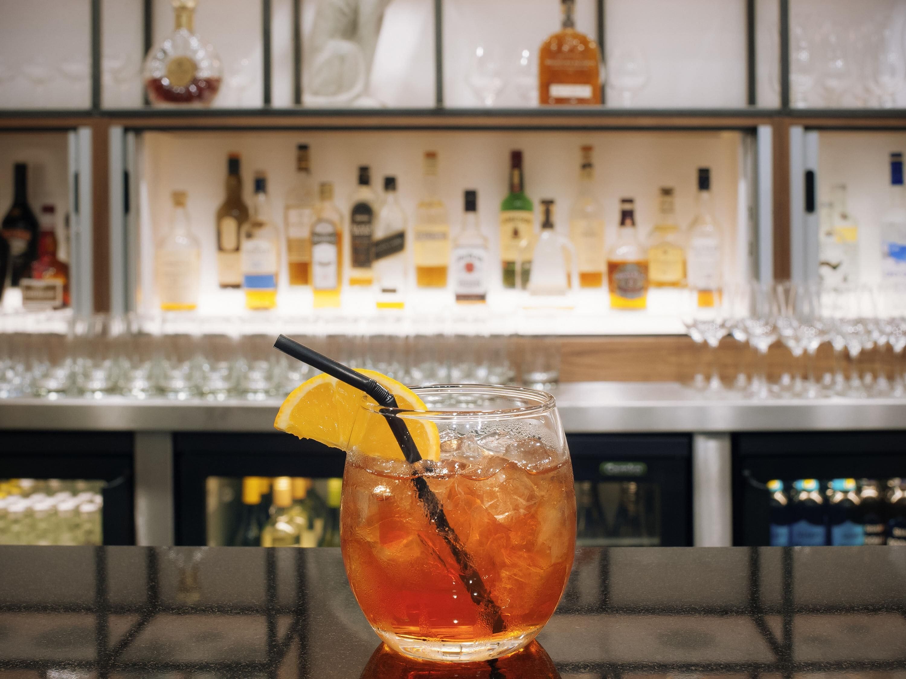 A bar setting with a glass of amber-colored drink garnished with an orange slice and a black straw. Behind the bar, there are shelves stocked with various bottles of alcohol.