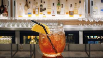 A bar setting with a glass of amber-colored drink garnished with an orange slice and a black straw. Behind the bar, there are shelves stocked with various bottles of alcohol.