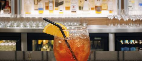 A bar setting with a glass of amber-colored drink garnished with an orange slice and a black straw. Behind the bar, there are shelves stocked with various bottles of alcohol.