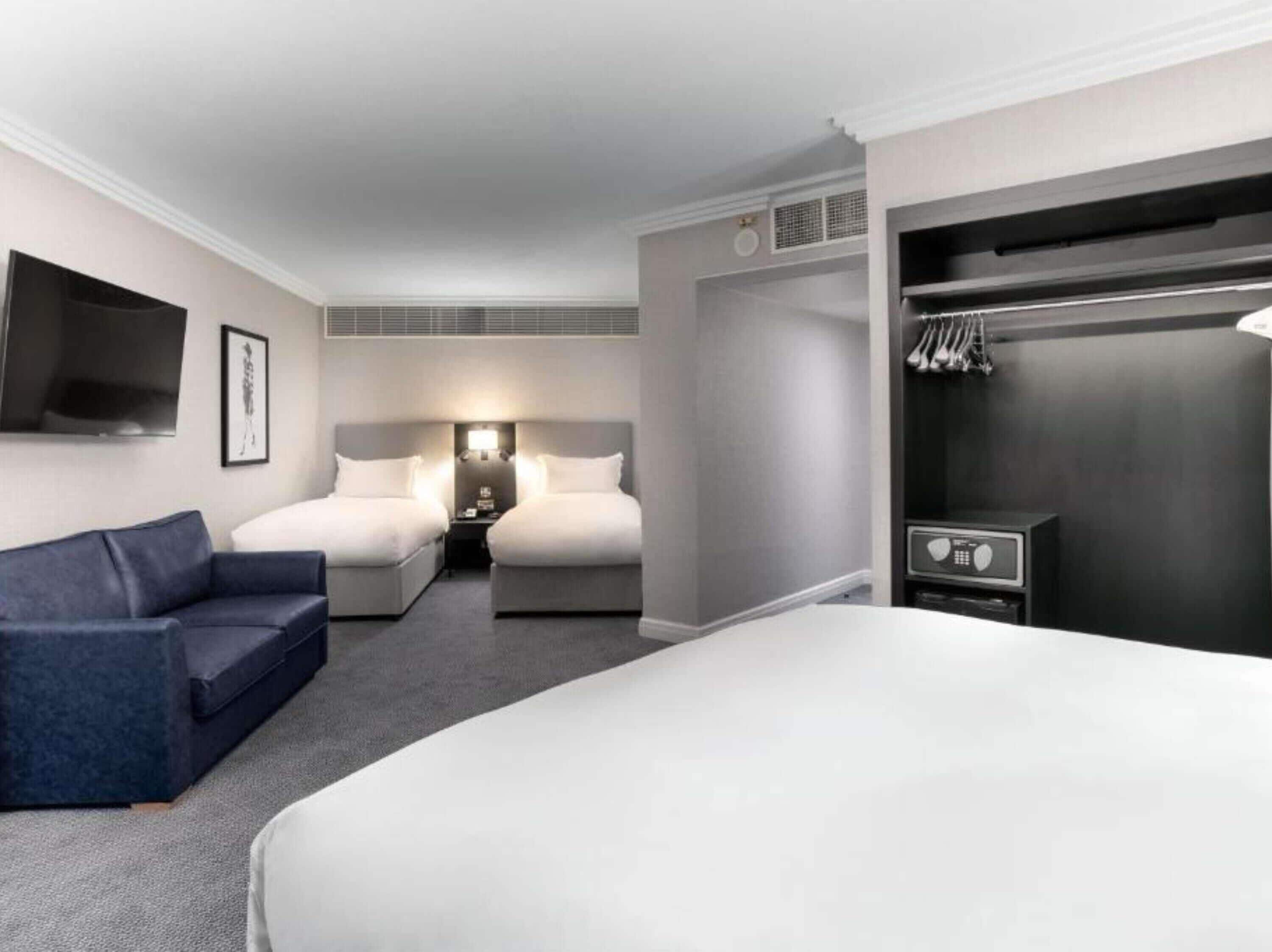 A hotel room with a large bed, a smaller bed, a sofa, a TV, a wardrobe, and a bedside table with a lamp.